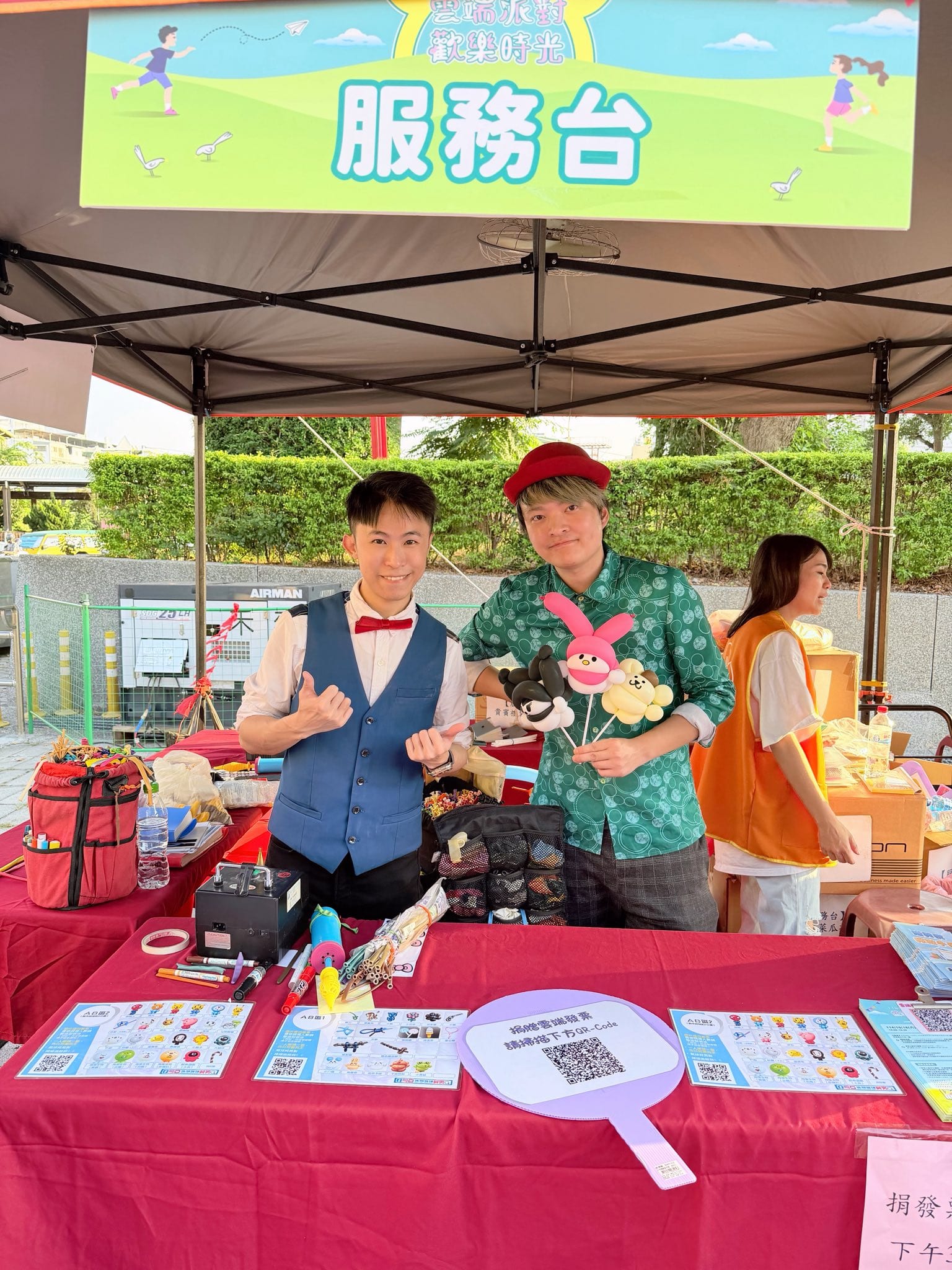 活動服務台帳篷下，柯男和戴紅帽的夥伴站在擺滿氣球用品的紅色桌子後方