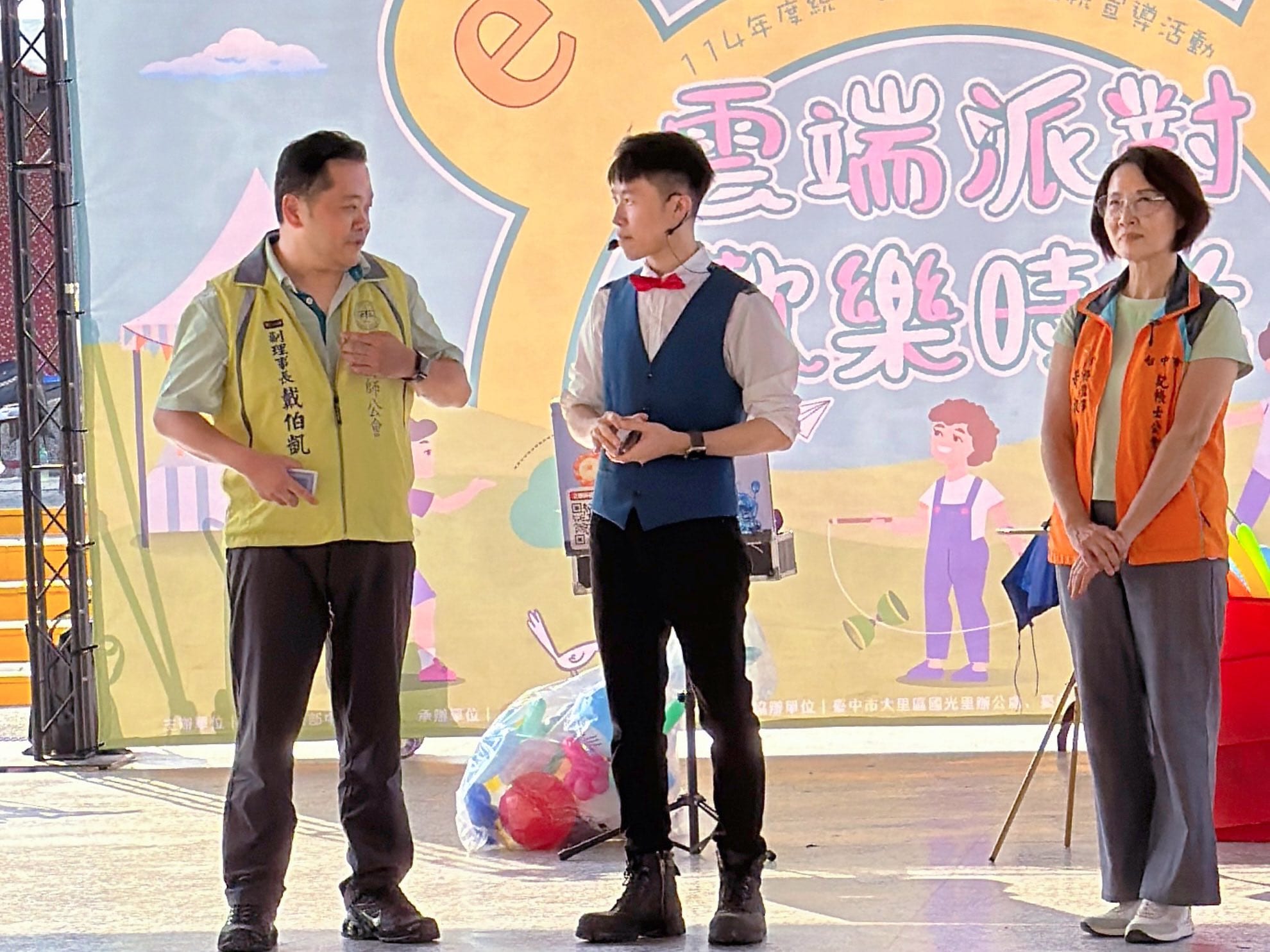 台中大里宣導活動舞台上，柯男站在兩位穿背心的活動人員中間，背後是雲端派對歡樂時光主視覺