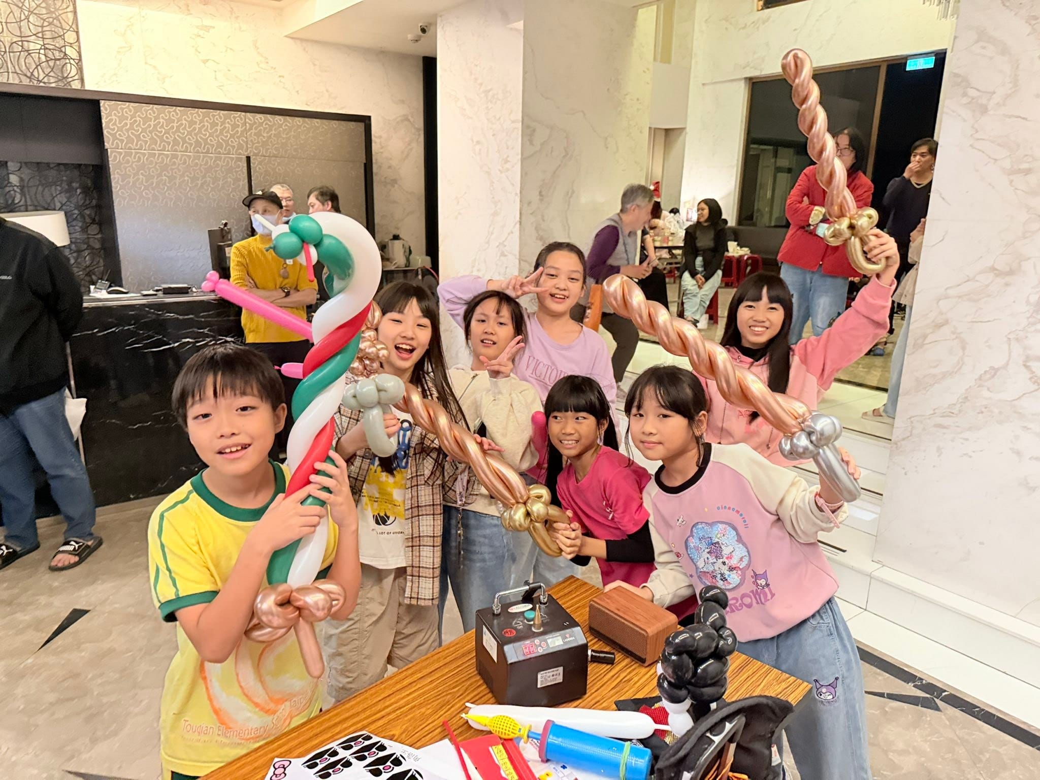 社區大廳裡一群小朋友舉著紅白綠、金色、銀色的寶劍造型氣球開心合照