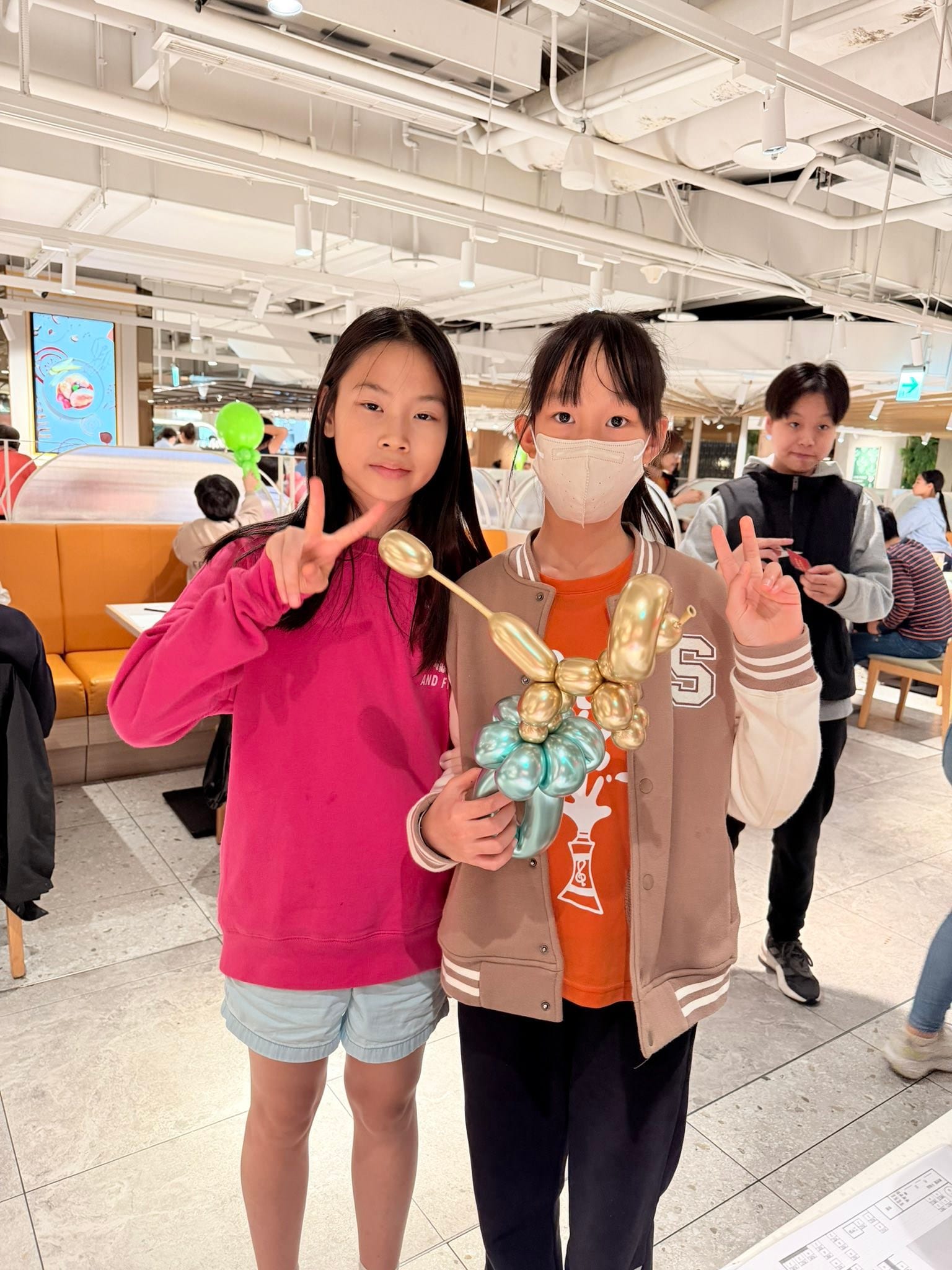 兩個女孩比 YA，其中一位拿著金色與藍綠金屬色的小狗造型氣球