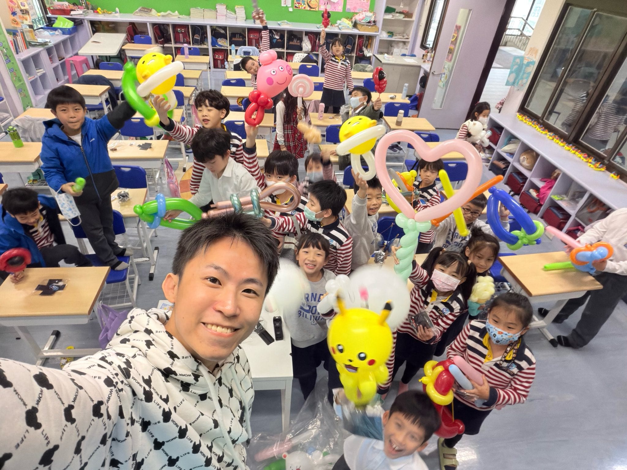 柯男在教室自拍，後方學生高舉皮卡丘、佩佩豬、棒棒糖和粉紅愛心造型氣球