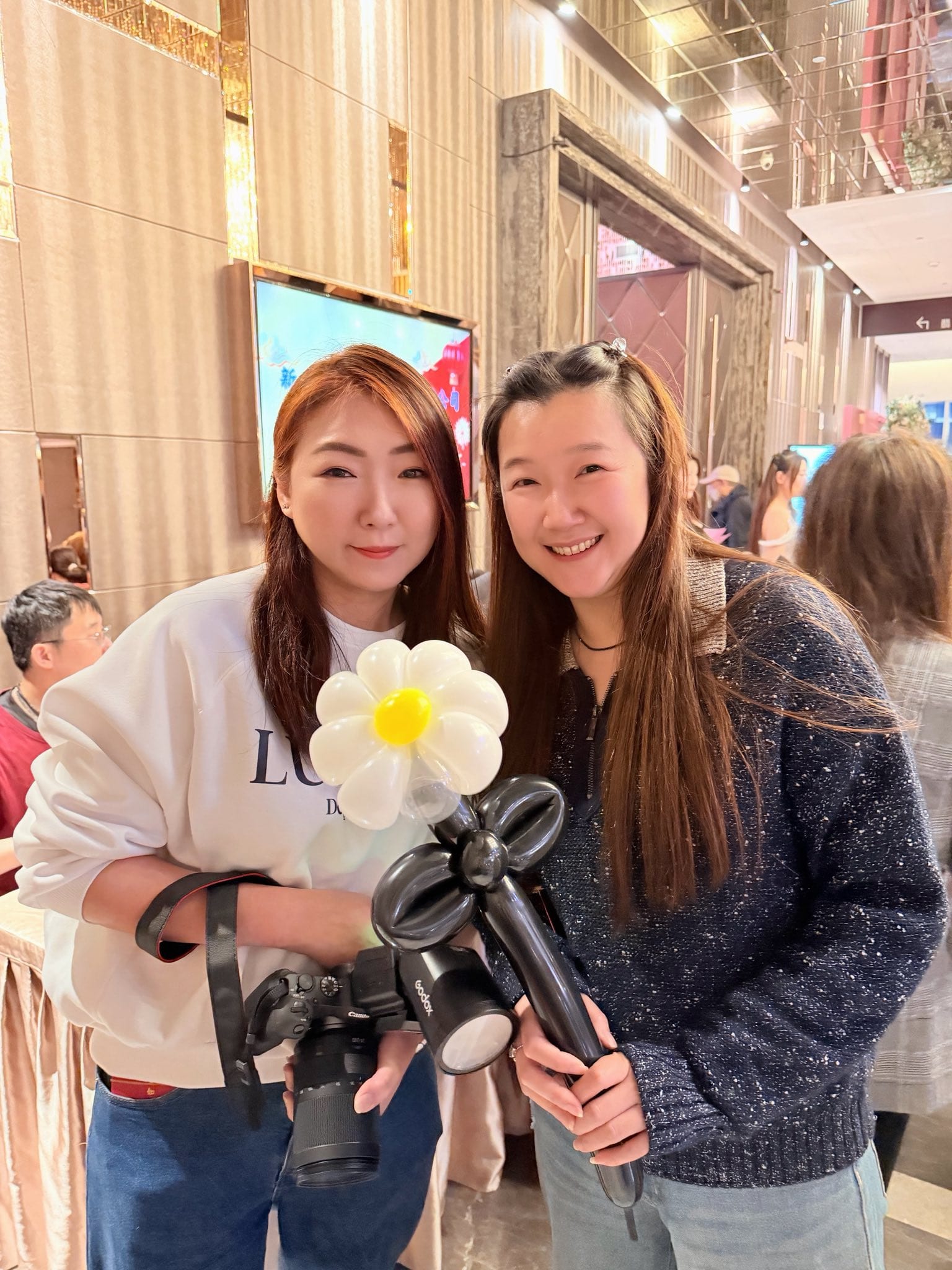 兩位女性來賓在宴會廳走道合照，其中一位拿著小雛菊造型氣球，另一位背著相機