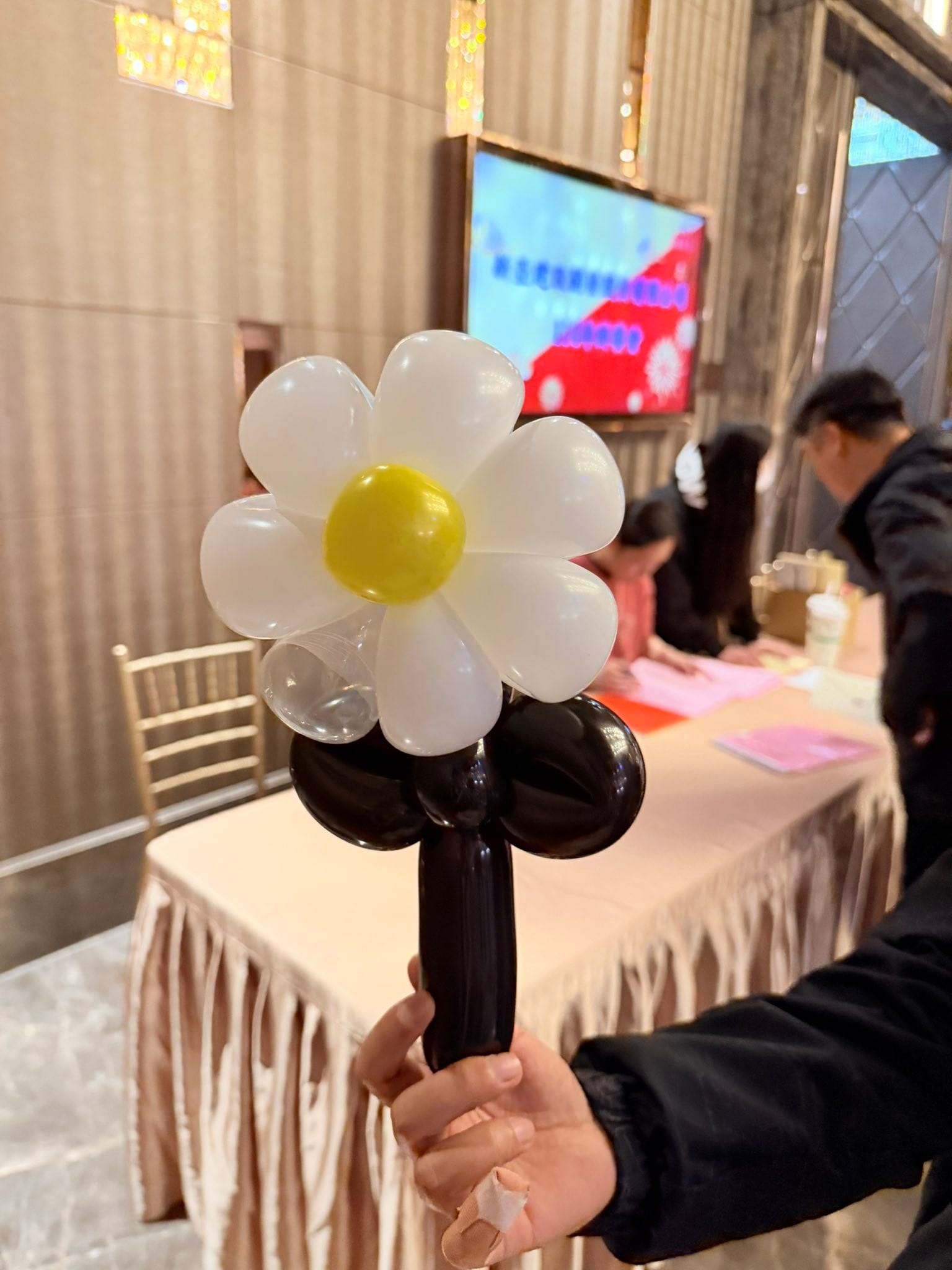 來賓手上拿著白色花瓣、黃色花心、黑色花莖的小雛菊造型氣球，背景是尾牙宴會廳報到桌