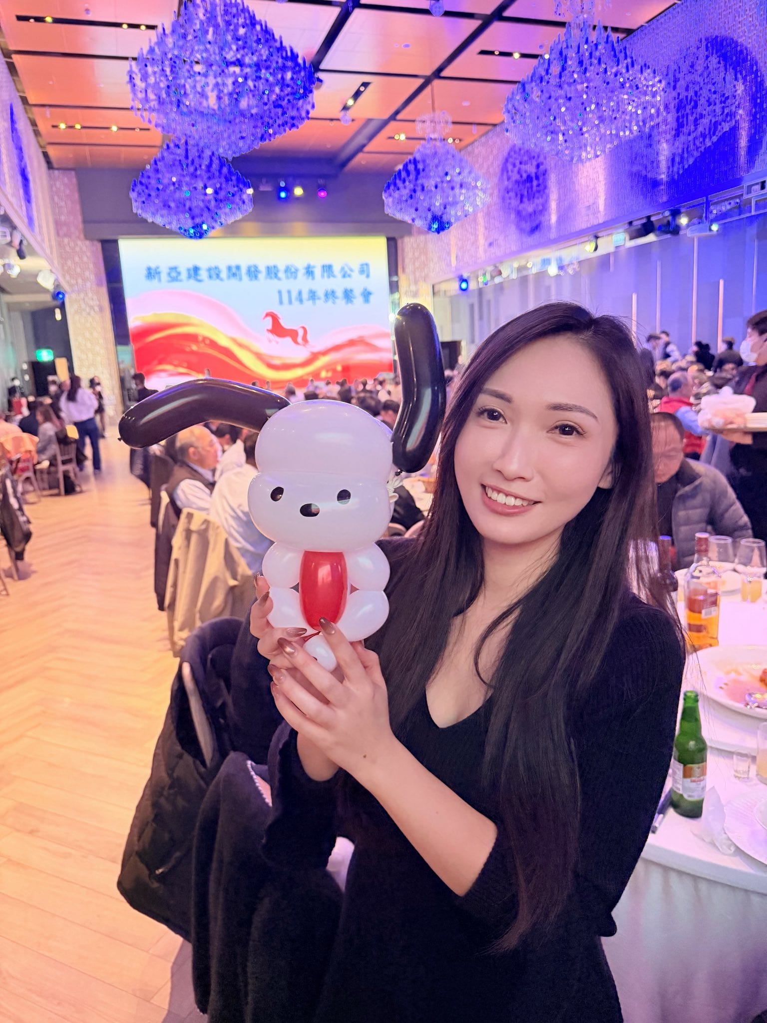 長髮女性在尾牙圓桌旁捧著白色身體、黑色長耳朵的小狗造型氣球，背景是水晶吊燈