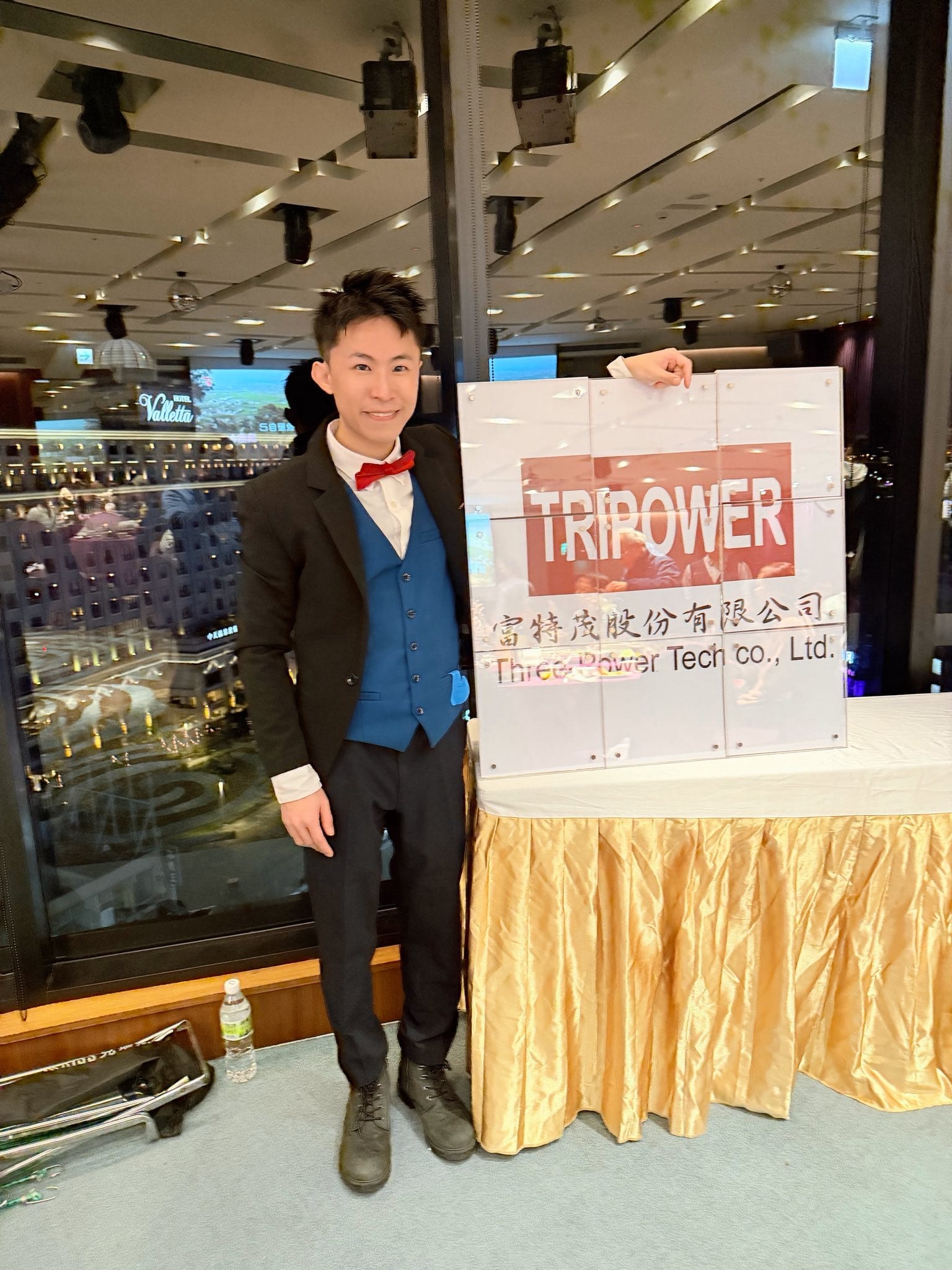 柯男穿黑色西裝外套、藍色背心與紅色領結,站在尾牙會場的白色看板旁合照