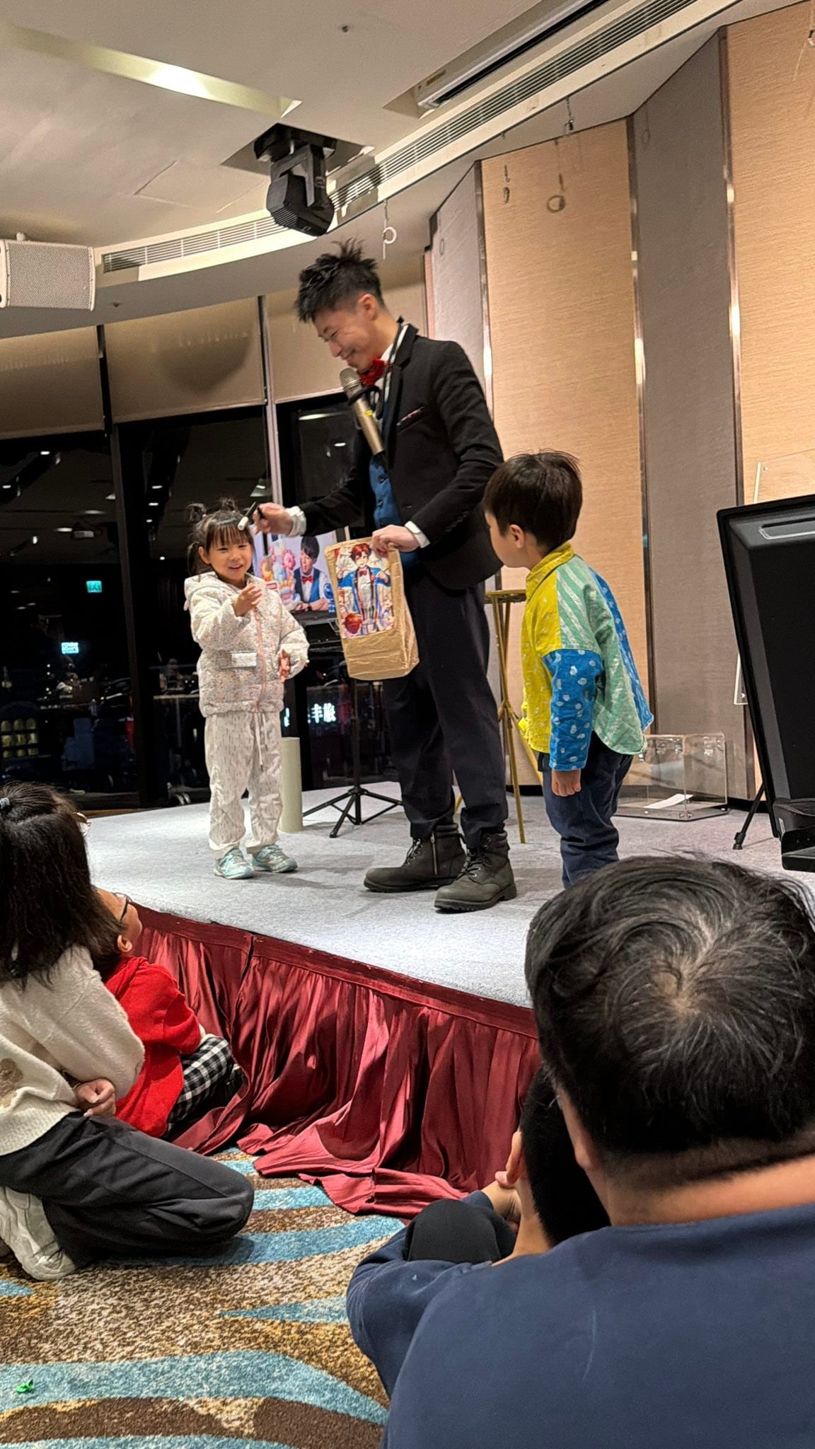 宜蘭礁溪尾牙舞台上,柯男拿著魔術道具紙袋,與穿白色連身衣的小女孩和穿彩色外套的男孩互動