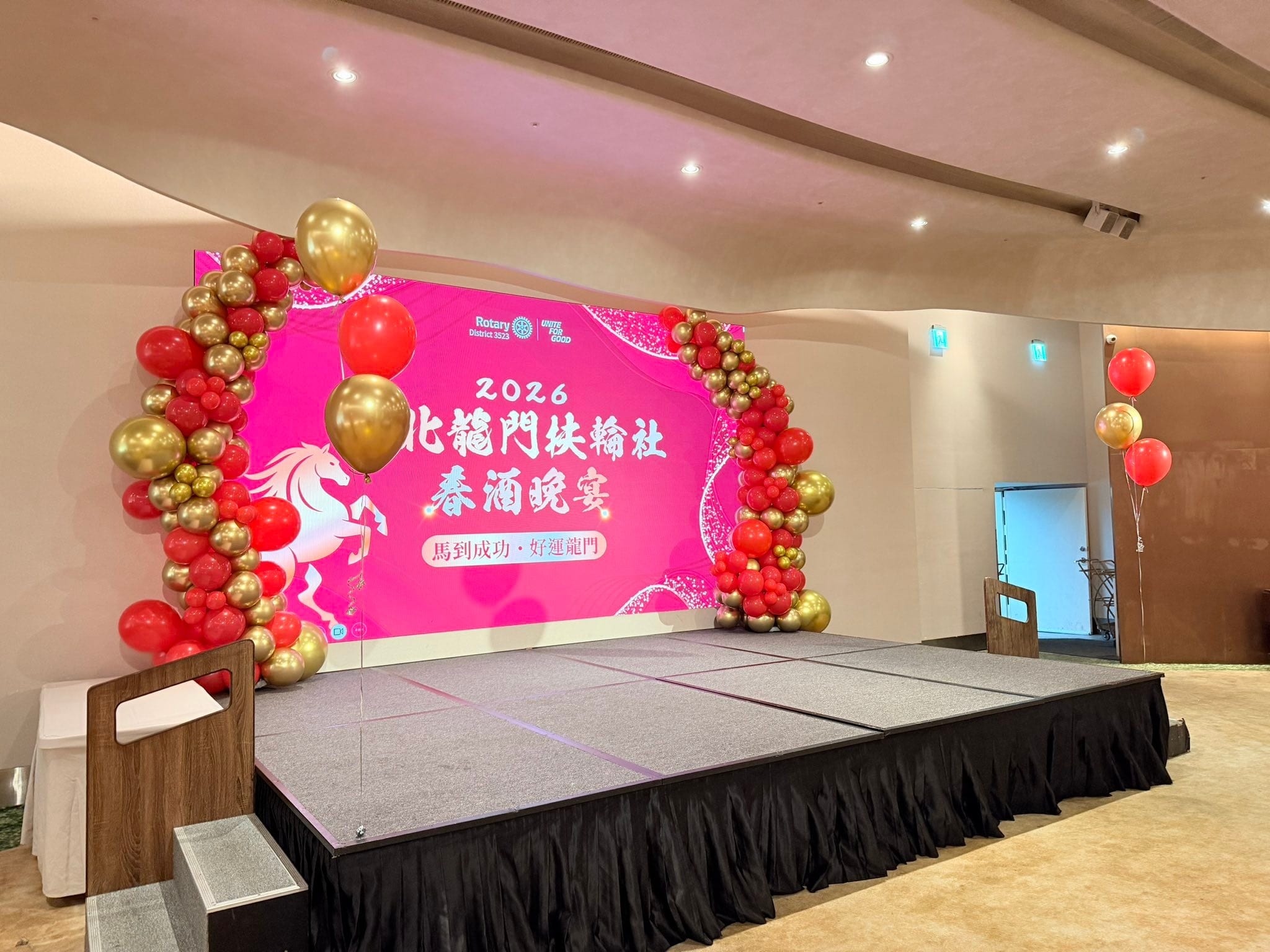 春酒晚宴舞台的粉紅背板兩側，以紅色與金色氣球做成不對稱的氣球拱門，旁邊有紅金空飄氣球