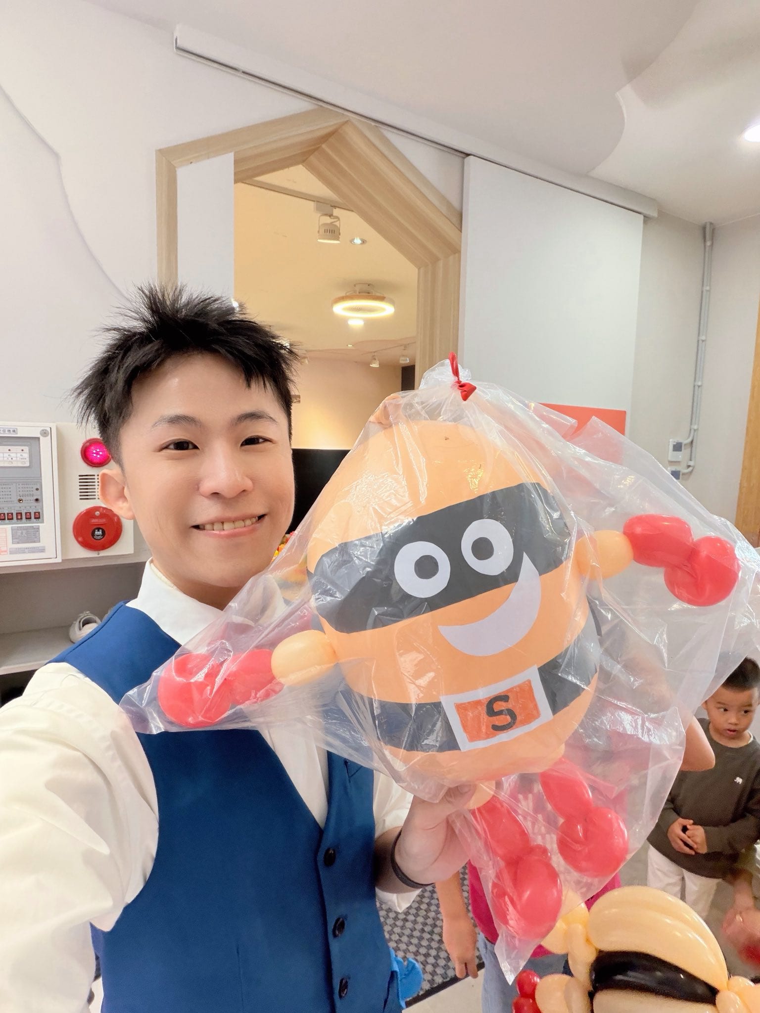 柯男在活動現場自拍，手上拿著裝進透明袋的土豆超人造型氣球，後方有小朋友