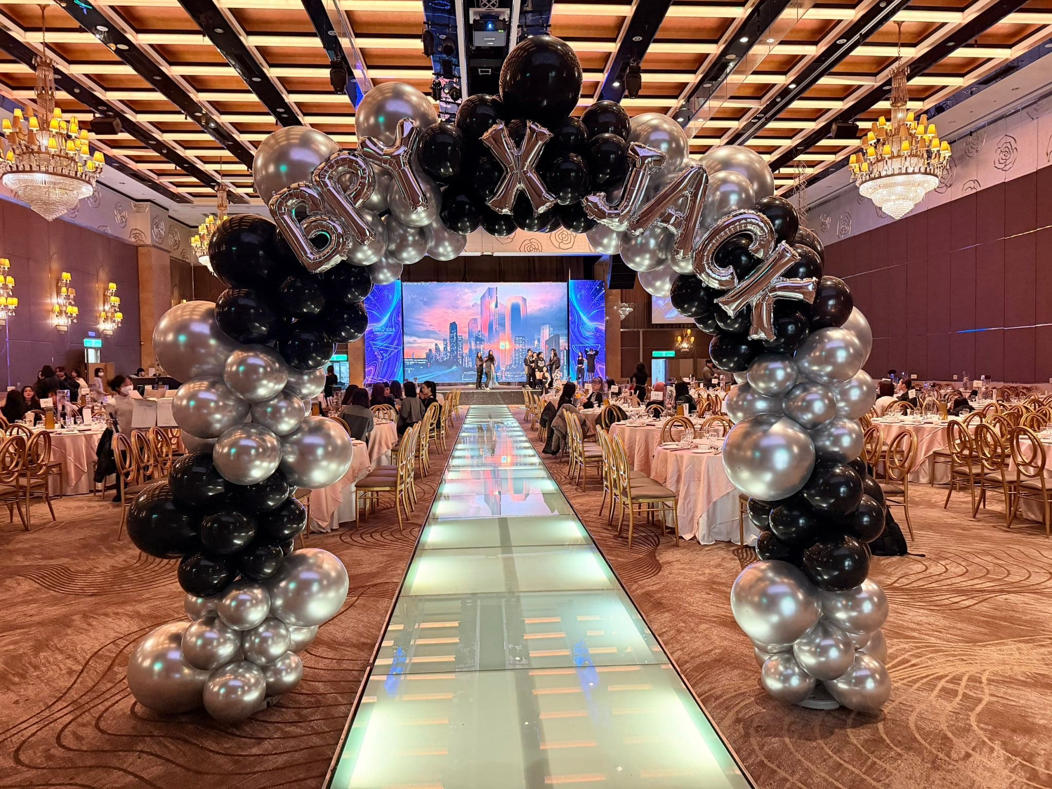 宴會廳伸展台入口的黑銀氣球拱門,拱門上方排著銀色鋁箔字母氣球,遠處是大型螢幕舞台