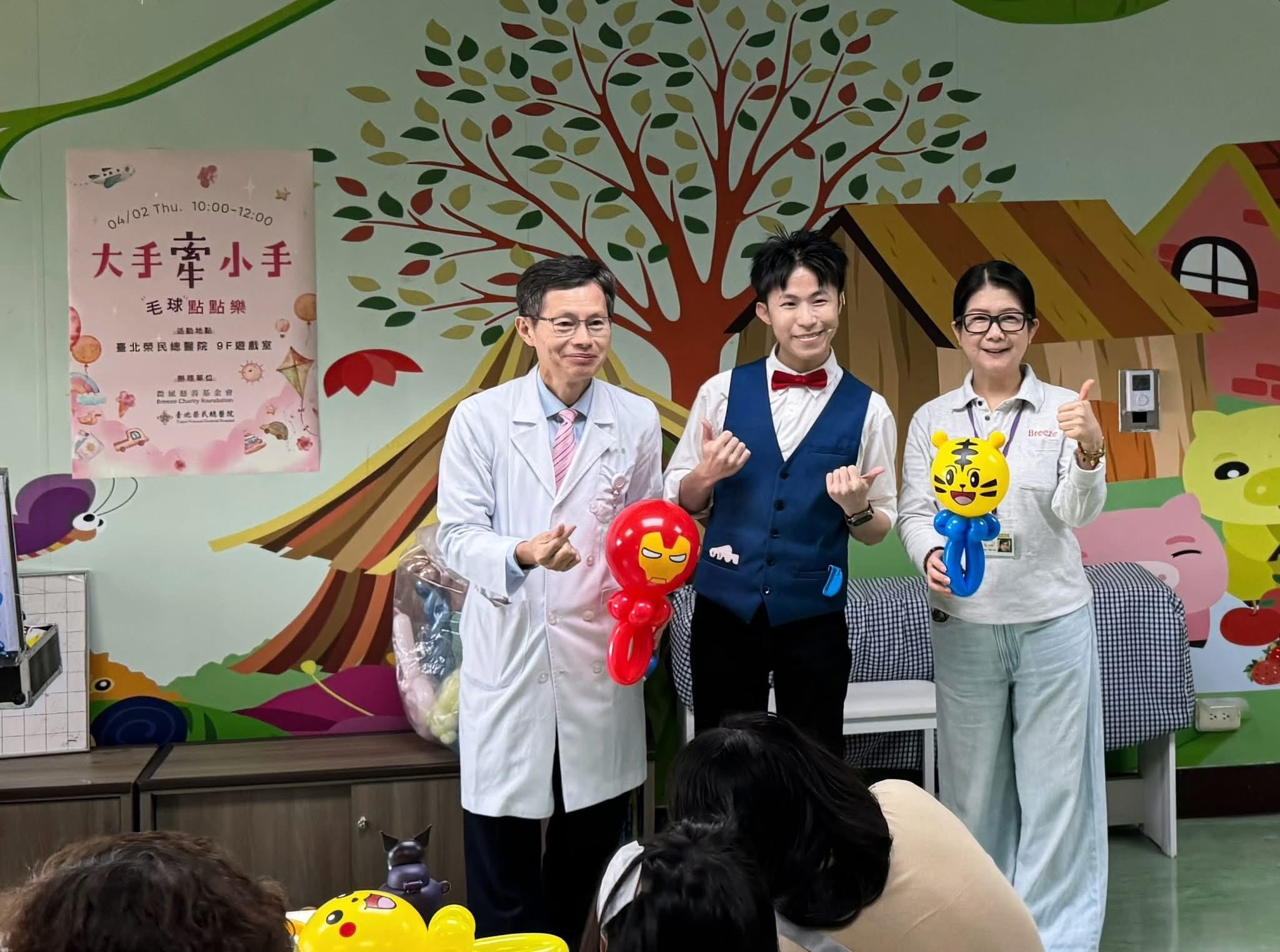 台北榮總兒童病房遊戲室，柯男與穿白袍的醫師、工作人員合照，兩人分別拿著鋼鐵人與巧虎造型氣球