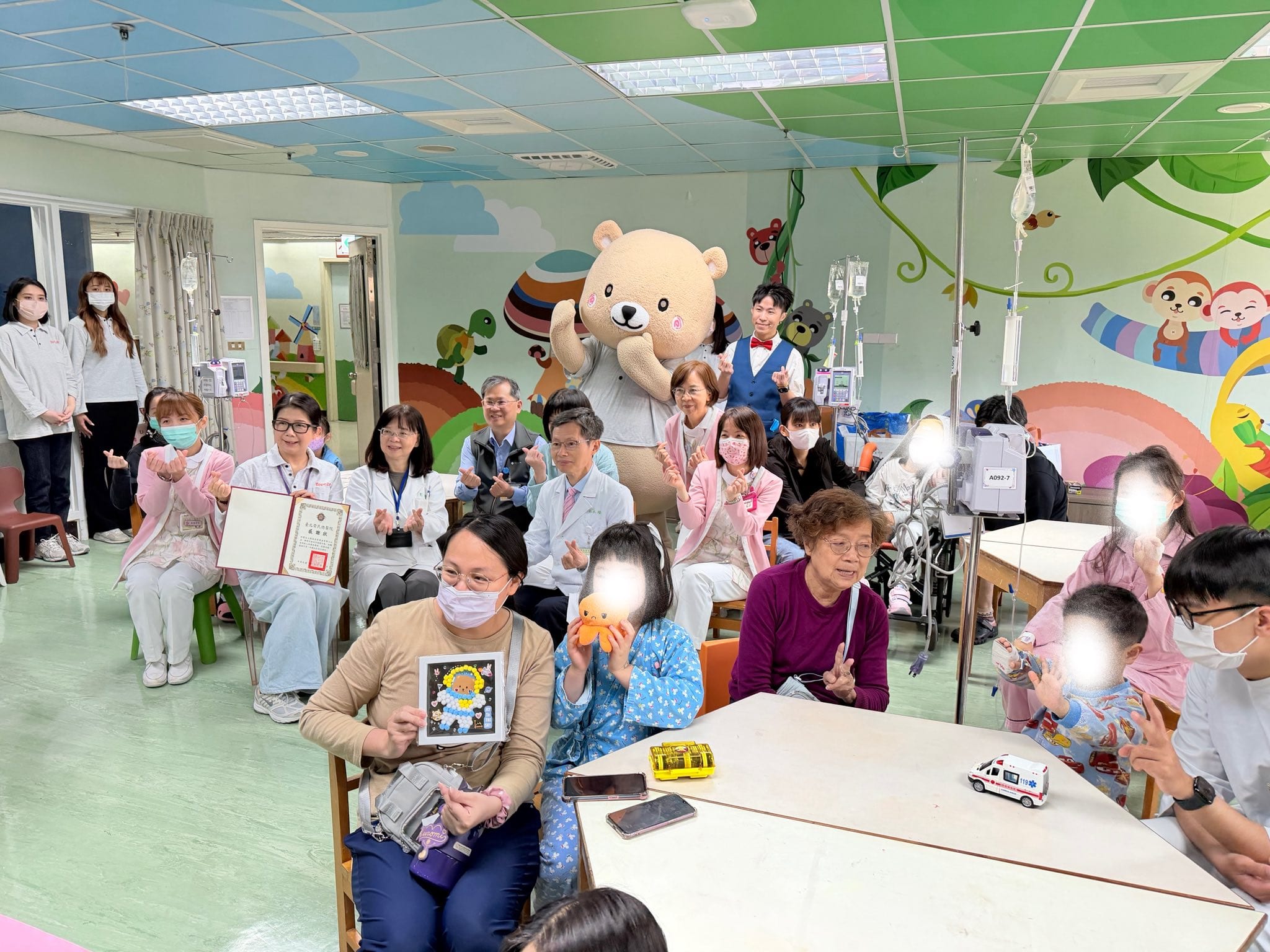 兒童病房遊戲室裡家長、病童與醫護人員圍坐鼓掌，後方有熊熊玩偶裝與點滴架
