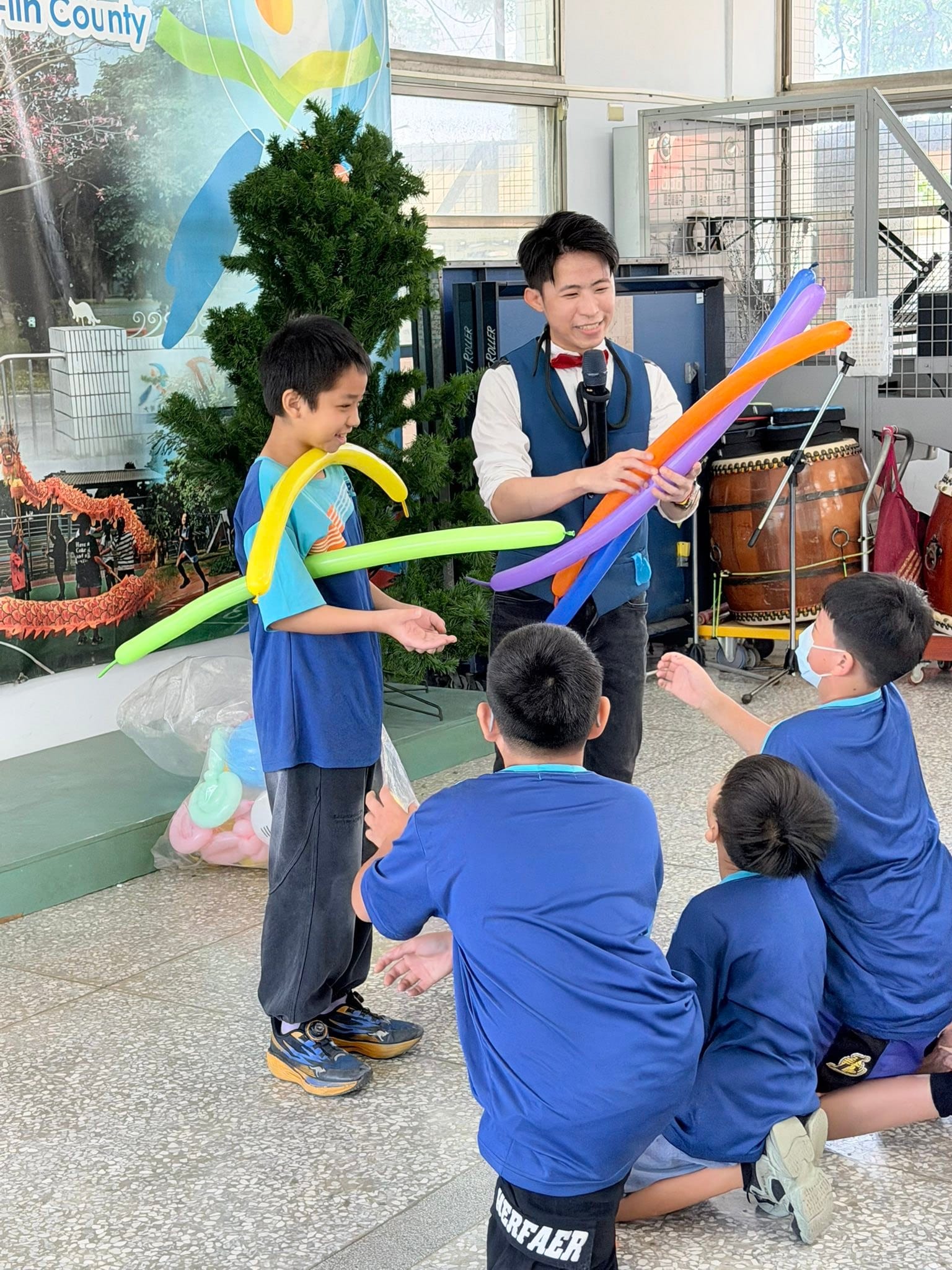 戴頭戴式麥克風的柯男把橘紫色長條氣球遞給上台的男孩,旁邊同學蹲坐觀看