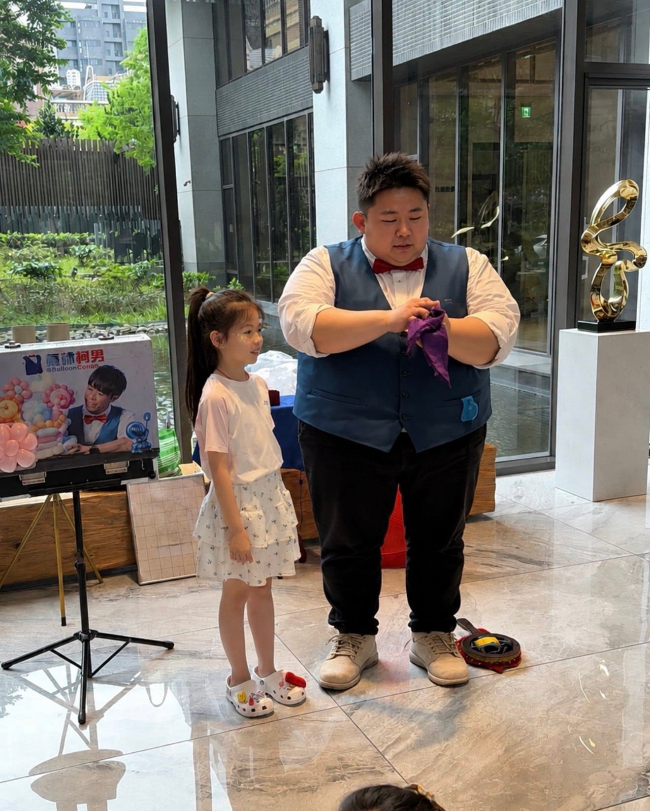 穿藍色背心的魔術師手拿紫色絲巾，身旁穿白色洋裝的小女孩上台協助表演