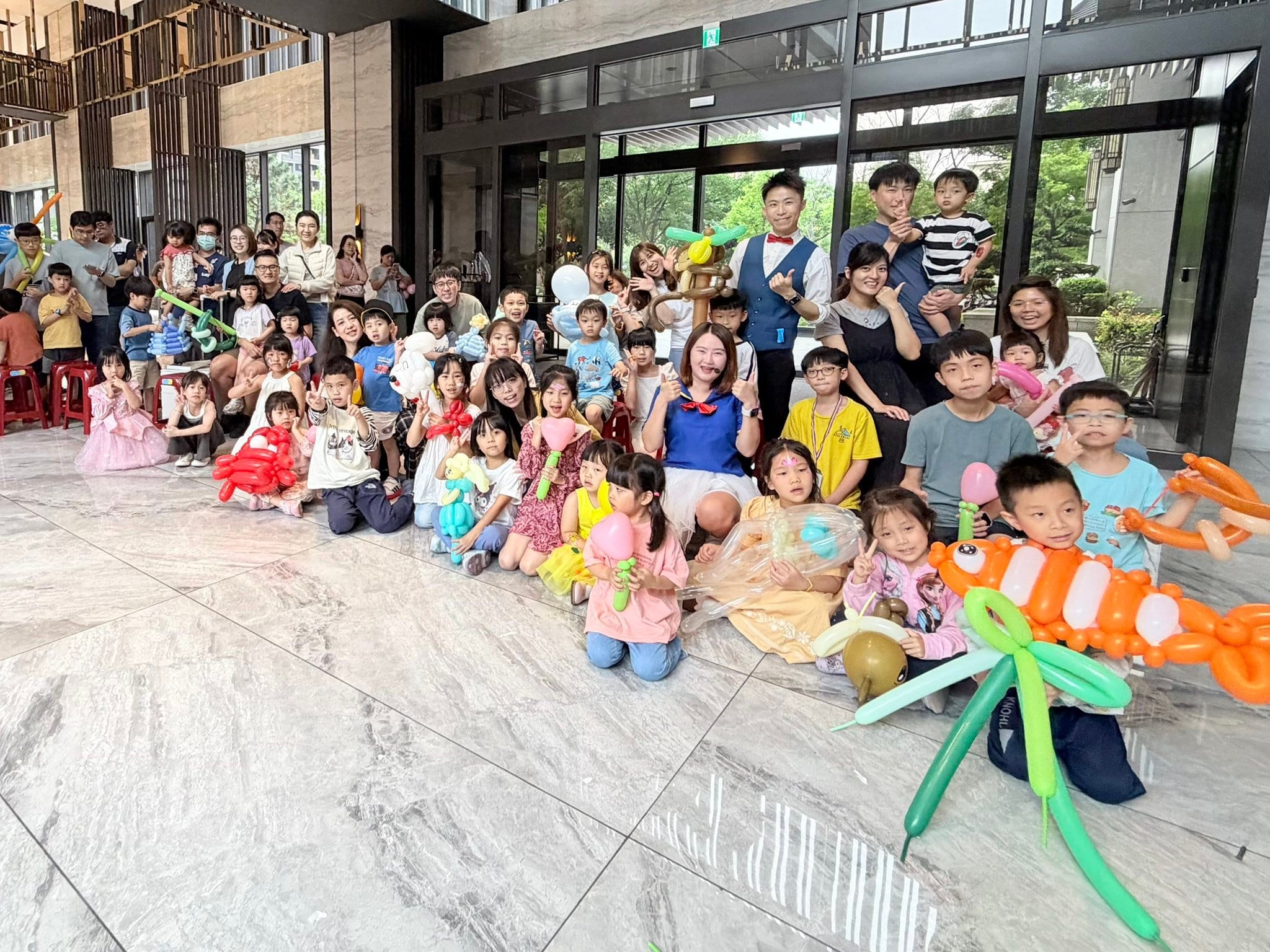 社區大廳內大人小孩一起合照，小朋友手拿橘色毛毛蟲、綠色等大型造型氣球