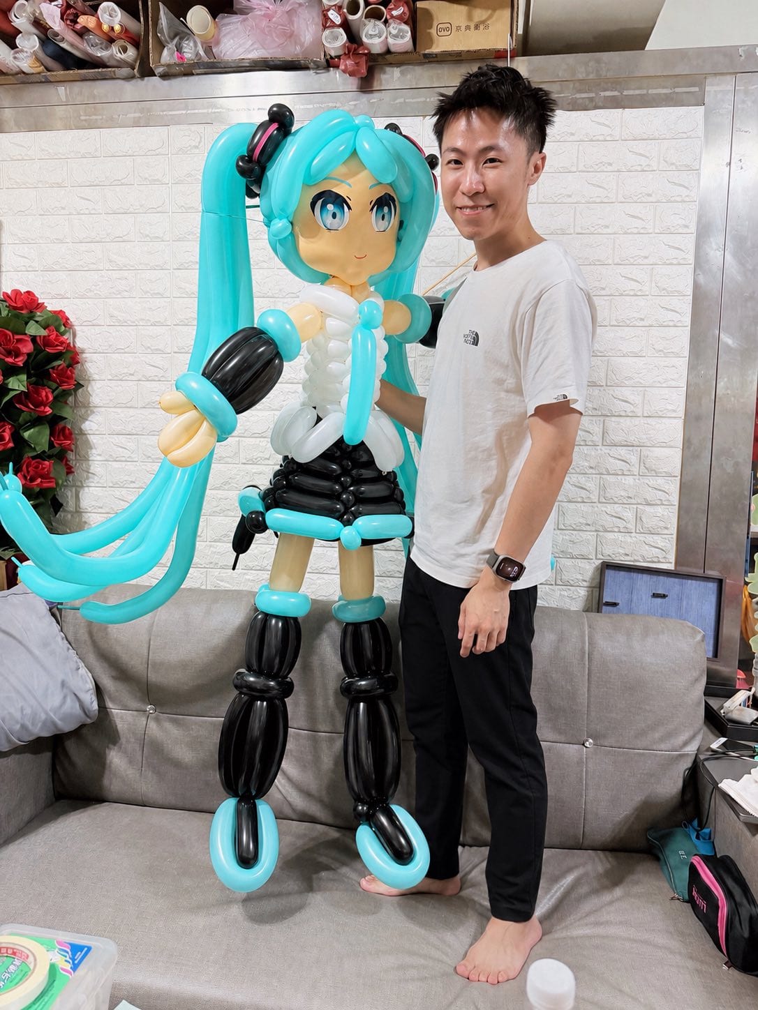 柯男站在與自己差不多高的初音未來氣球人偶旁,背景是白色磚牆與工作室沙發