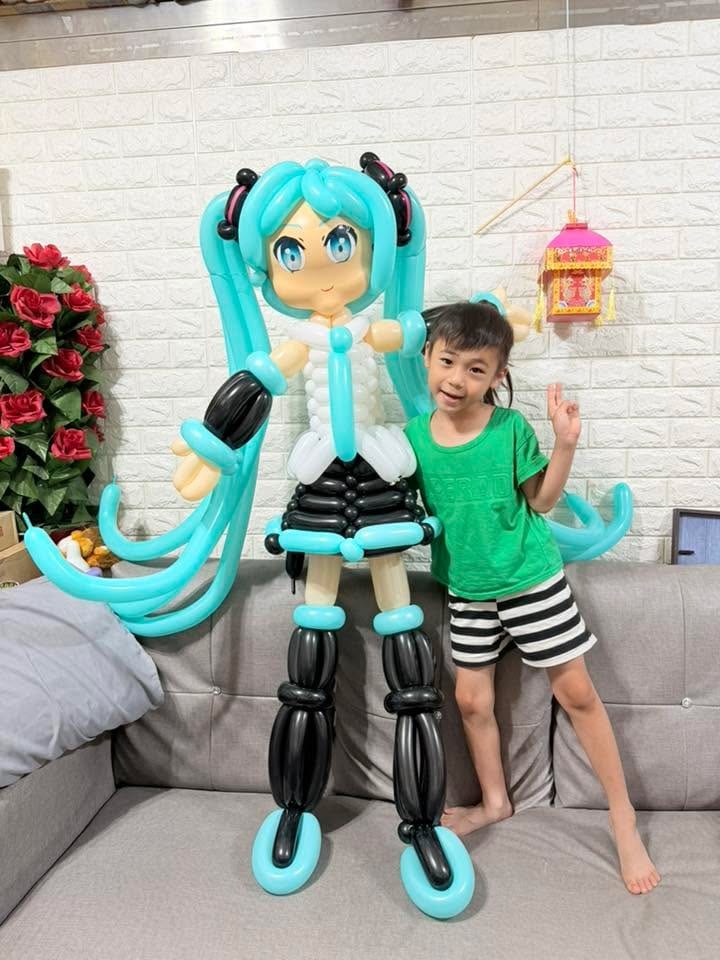 穿綠色上衣的小孩站在初音未來氣球人偶旁比 YA,人偶比孩子高出一截