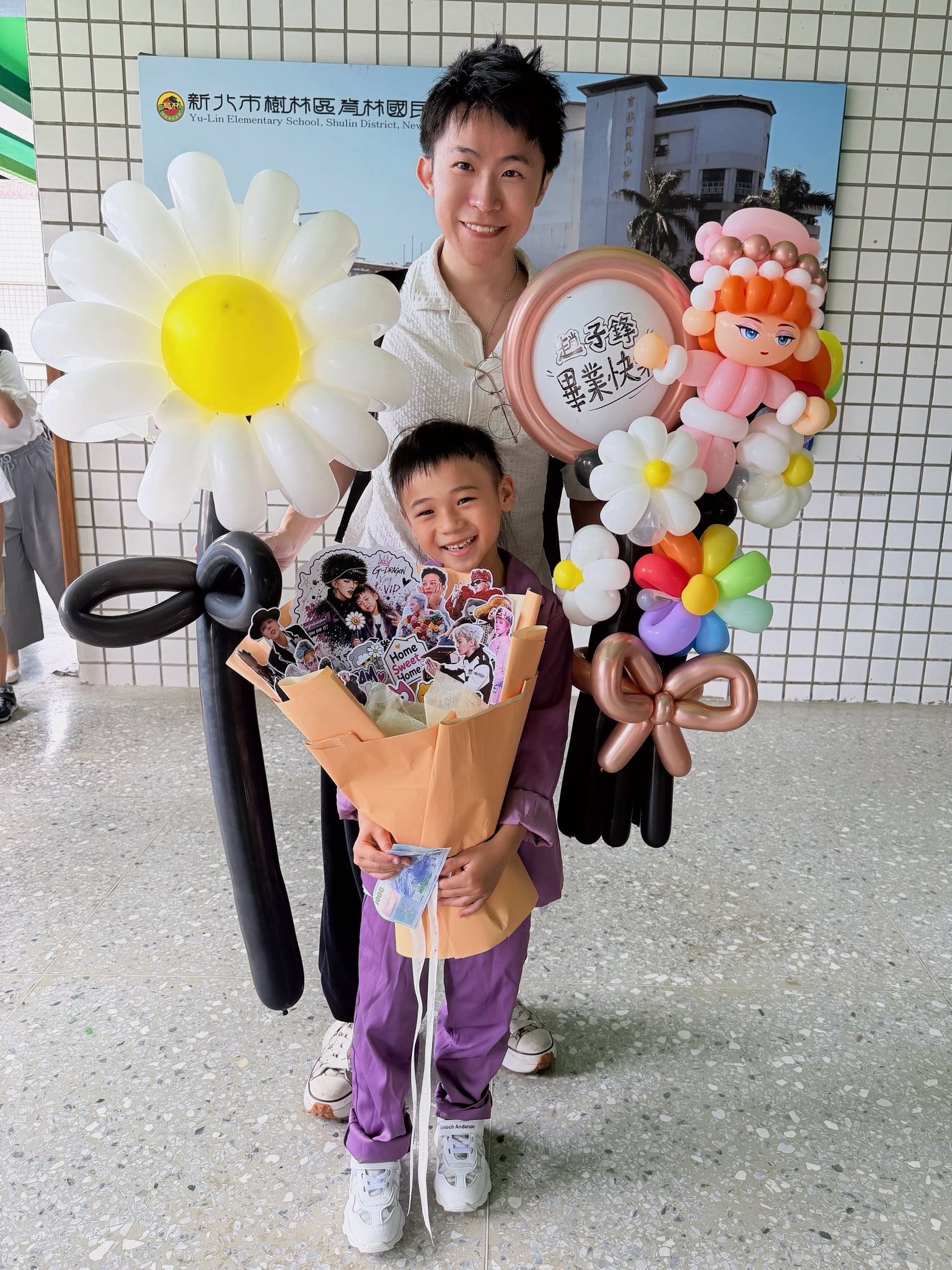 穿紫色畢業服的小男孩抱著 GD 卡片花束,身後柯男拿著白色小雛菊氣球花與粉紅人物氣球花束