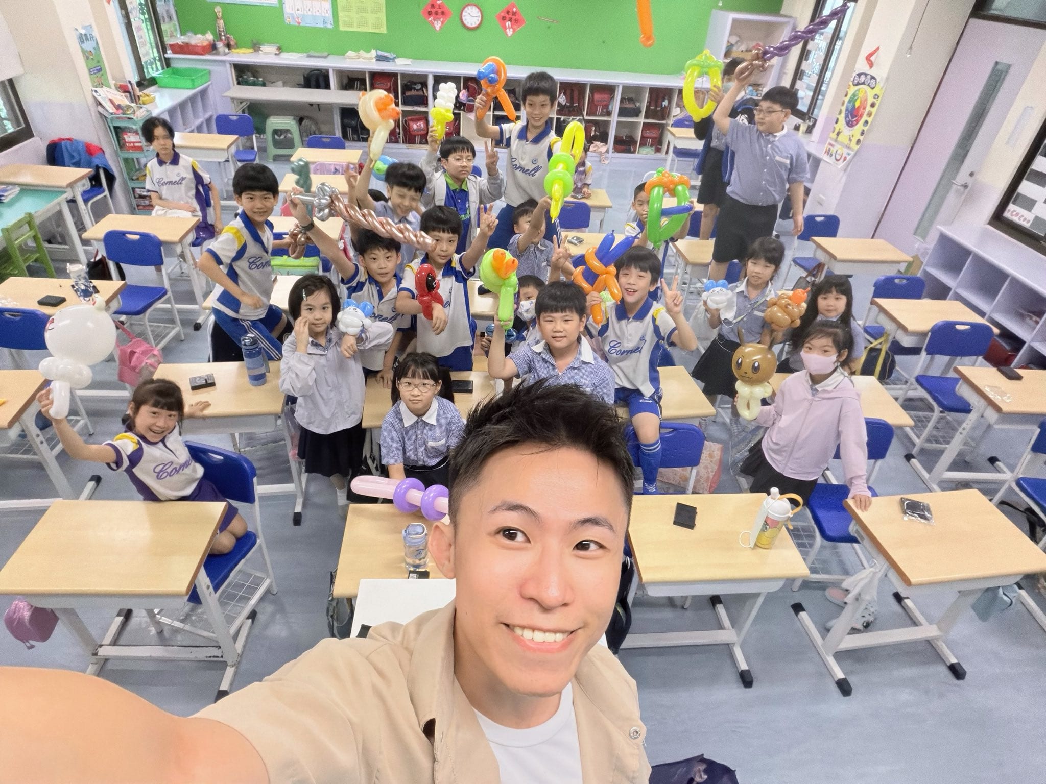 國小教室內學生站在課桌椅間比 YA，手上拿著小狗、寶劍、綠色造型等各式氣球