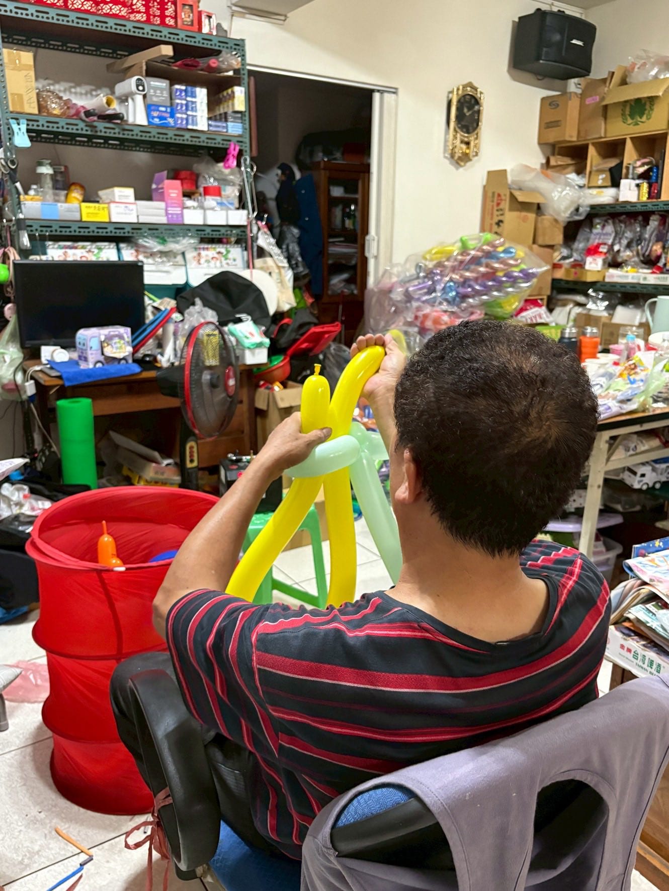 穿紅黑條紋上衣的男士背對鏡頭，坐在堆滿氣球與材料的房間裡折黃色造型氣球