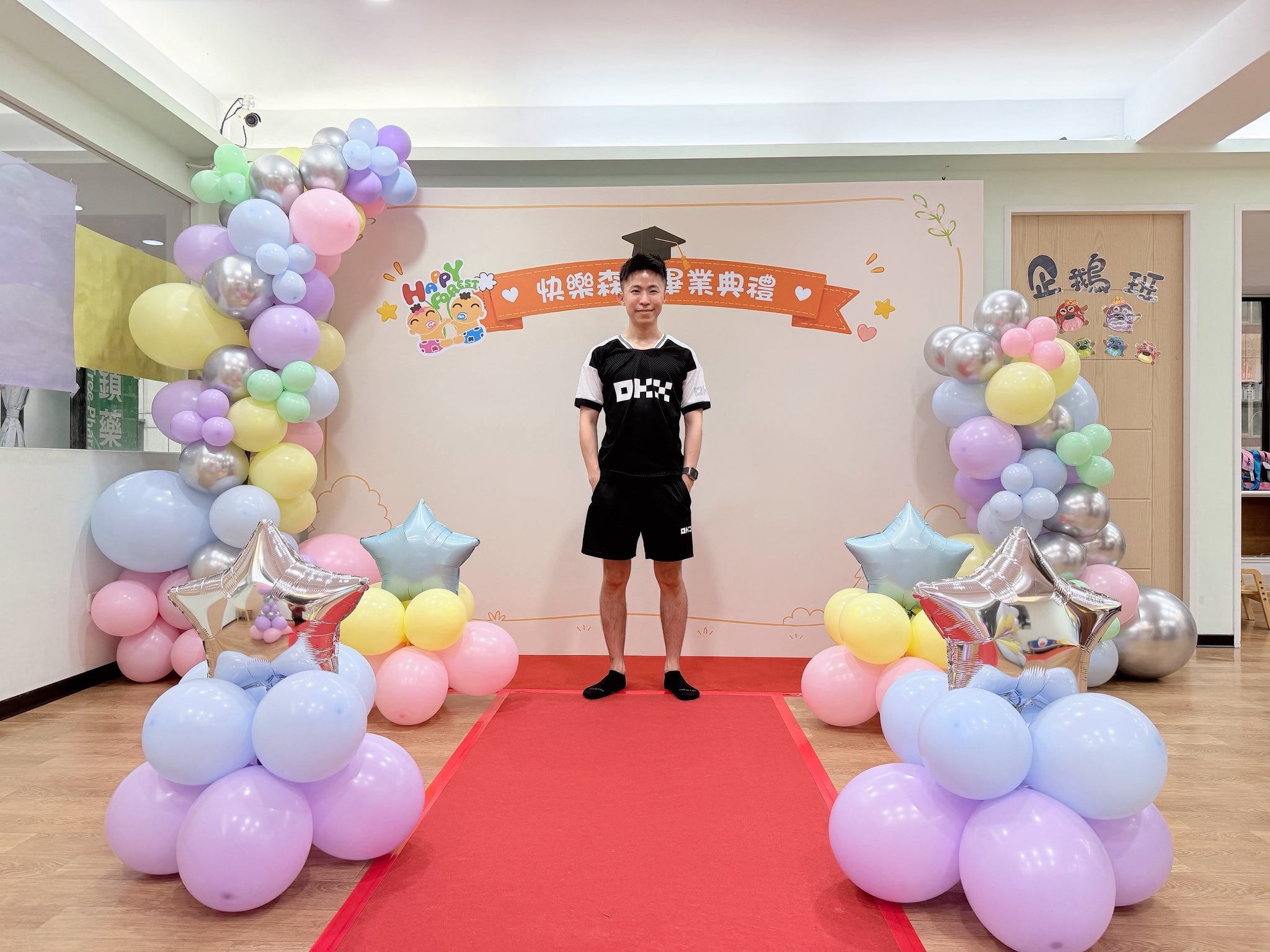 板橋快樂森林托嬰中心畢業典禮背板前鋪著紅地毯，兩側是馬卡龍色氣球串與銀色星星氣球，柯男站在中央