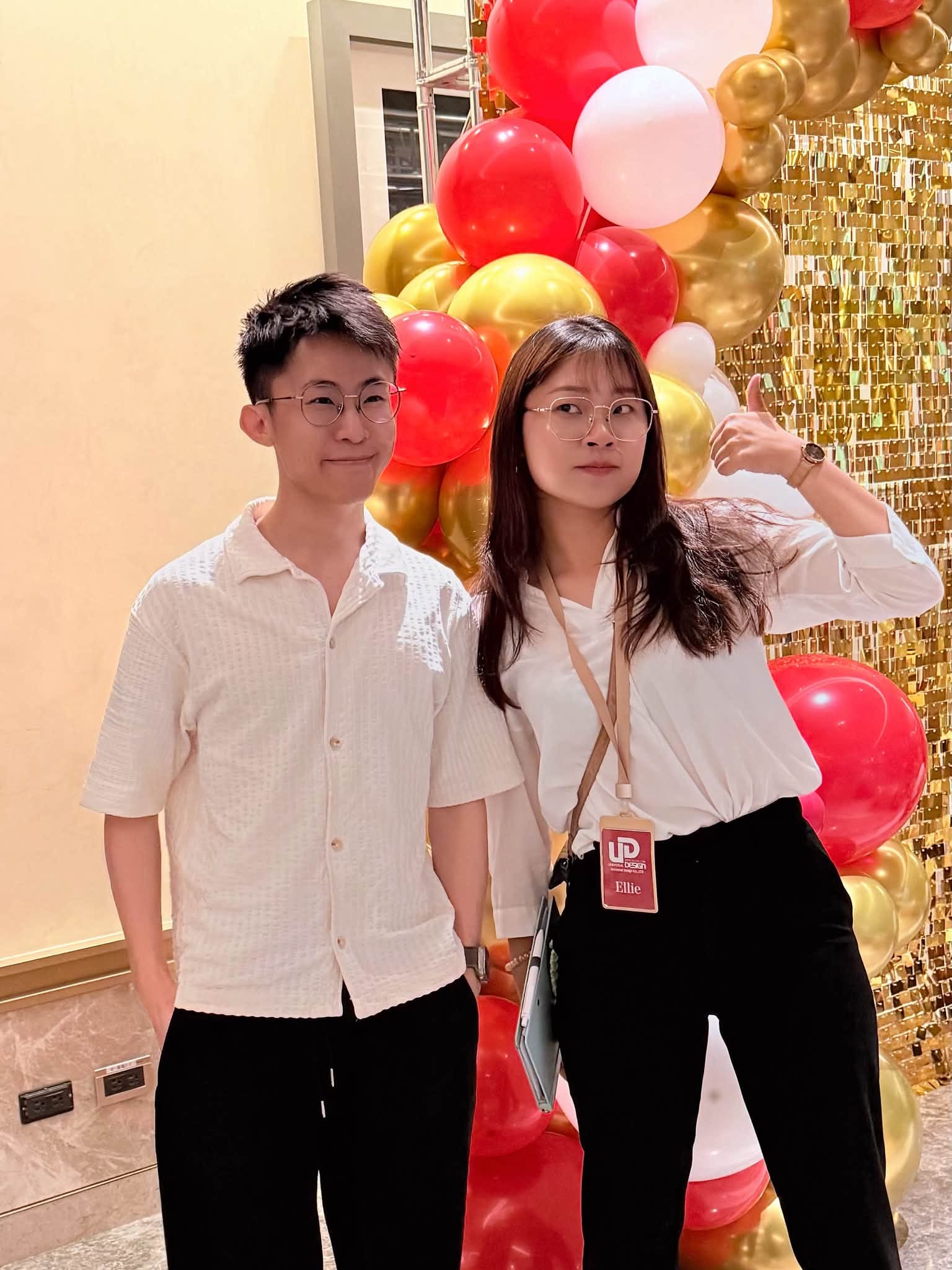 兩位穿白色上衣的年輕人站在紅金氣球串與金色亮片牆前合照，其中一人比讚