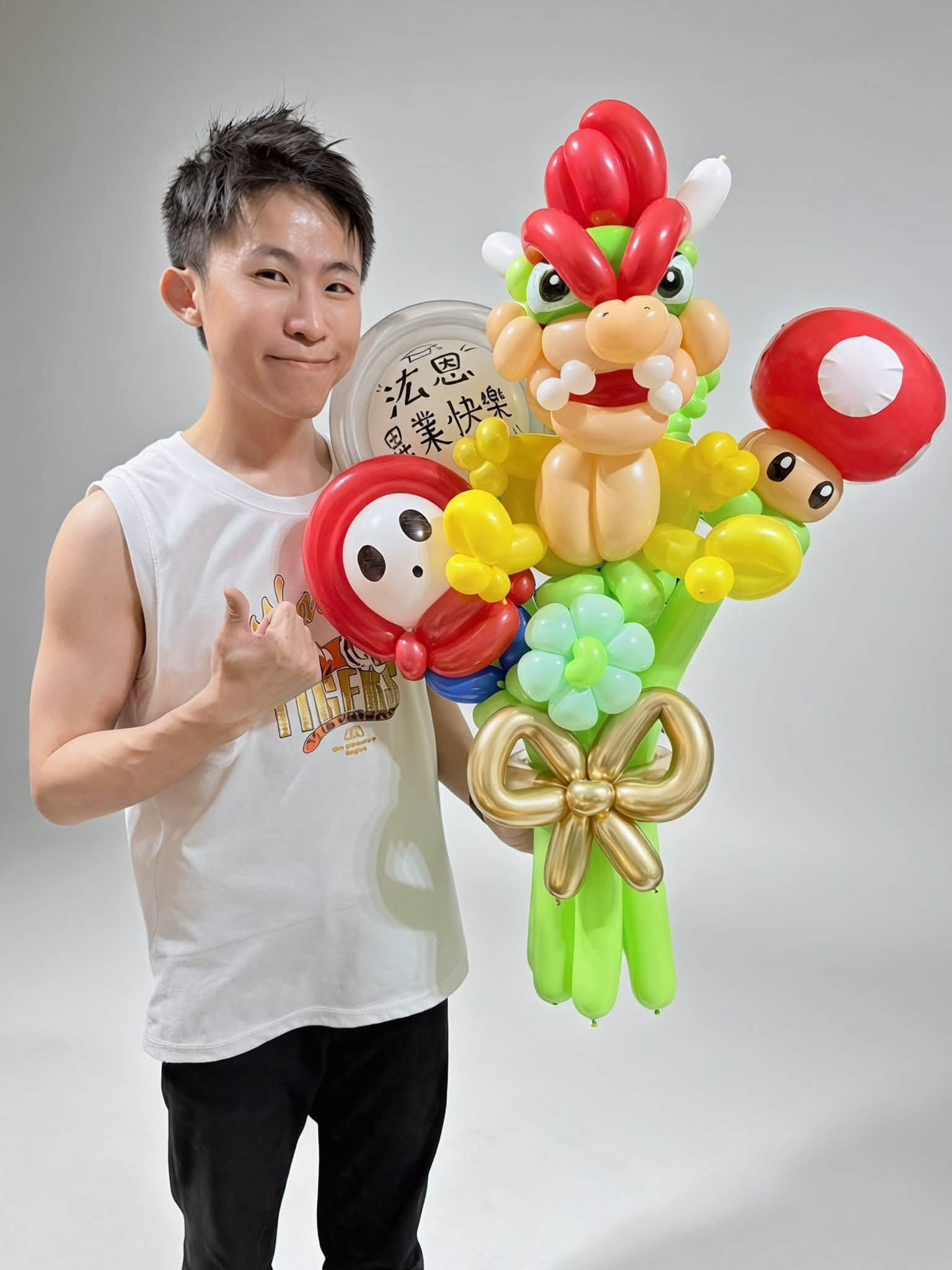 穿白色背心的柯男比讚，手抱庫巴氣球花束，花束後方圓框貼著畢業快樂客製文字