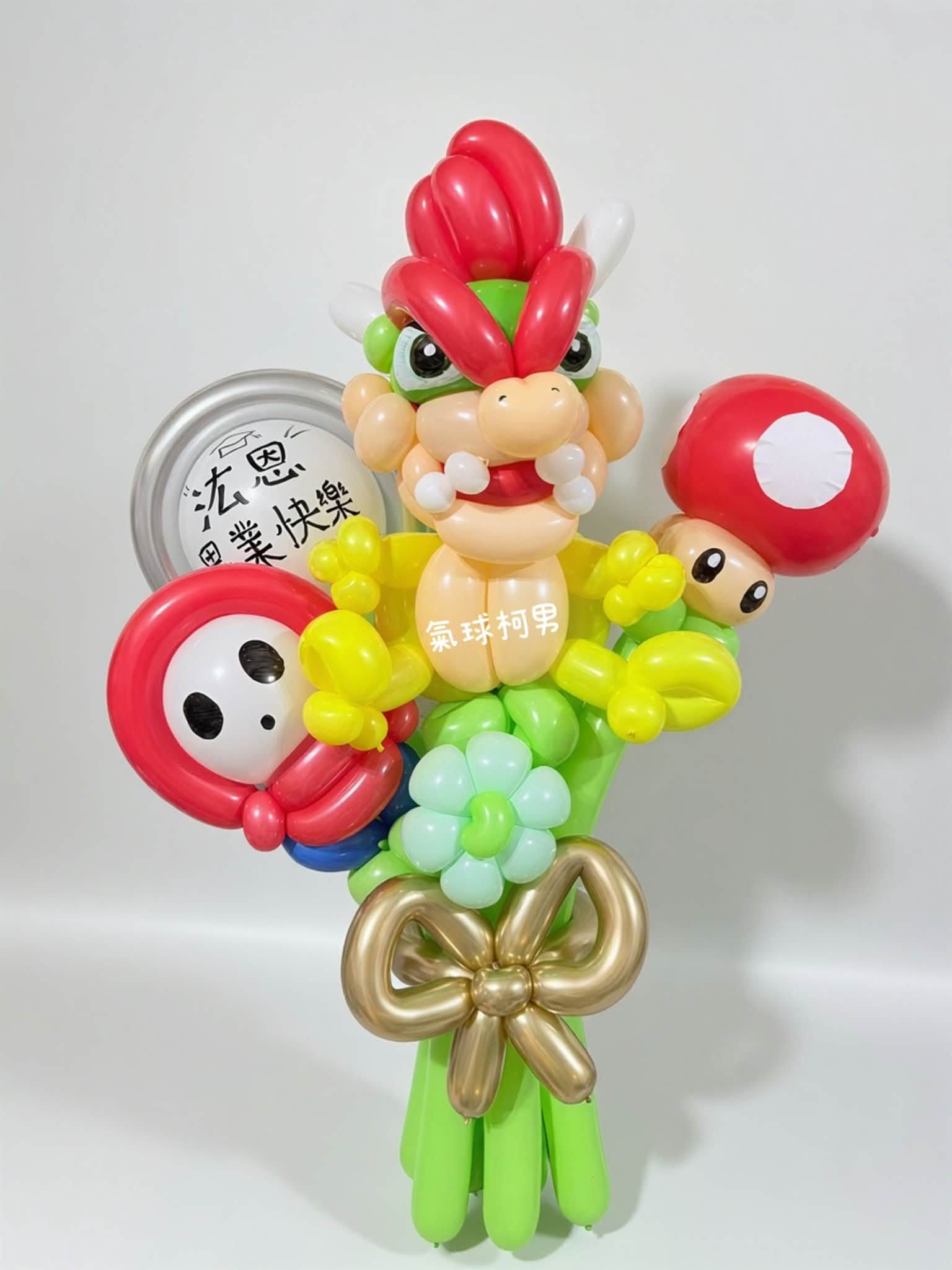 庫巴畢業氣球花束，紅色頭髮、綠色龜殼的庫巴居中，左邊紅色斗篷白面具的嘿呵，右邊紅白點超級蘑菇，下方金色蝴蝶結