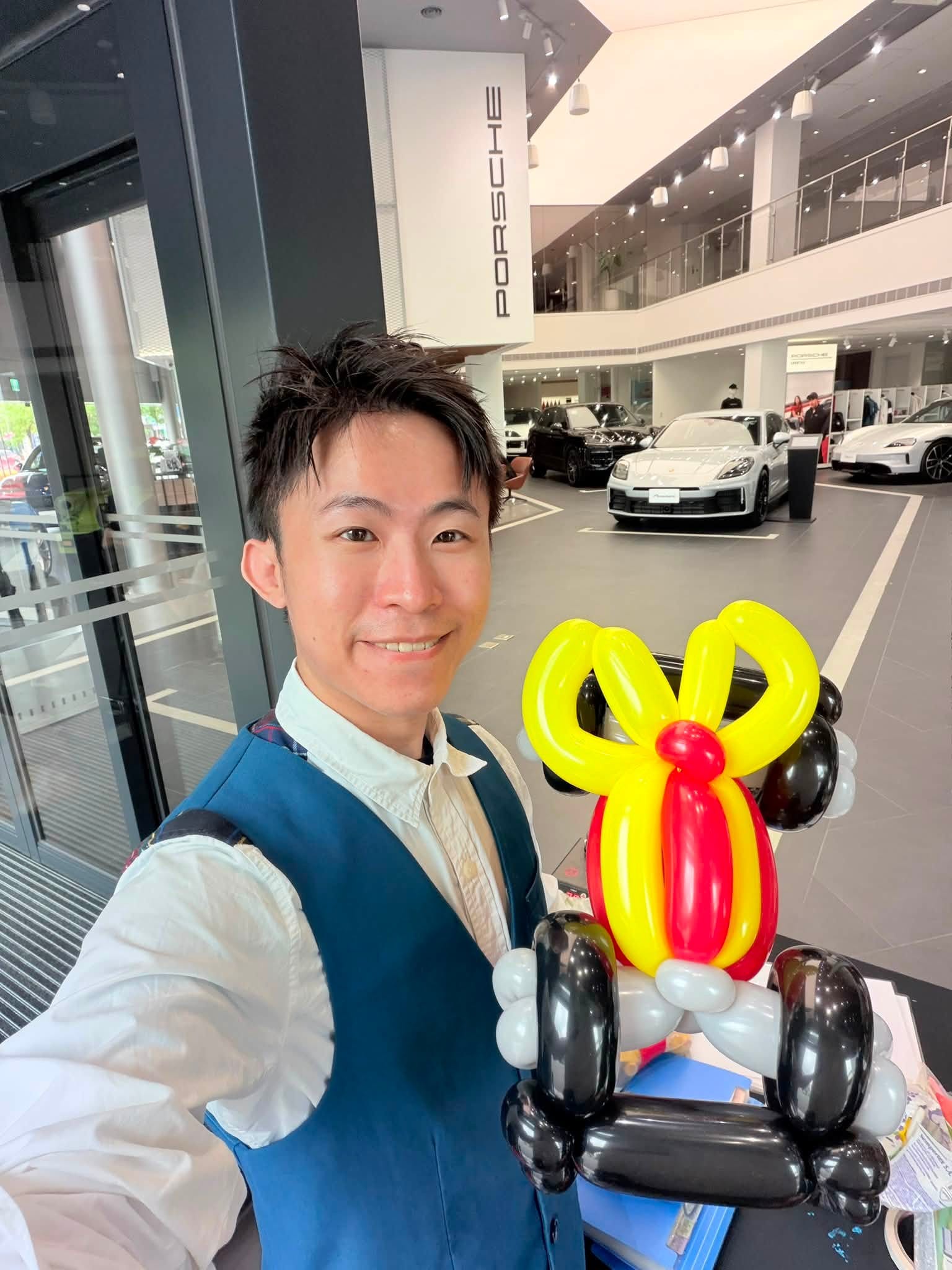 柯男手拿紅黃黑三色車子造型氣球自拍,背景是 PORSCHE 招牌與白色展示車