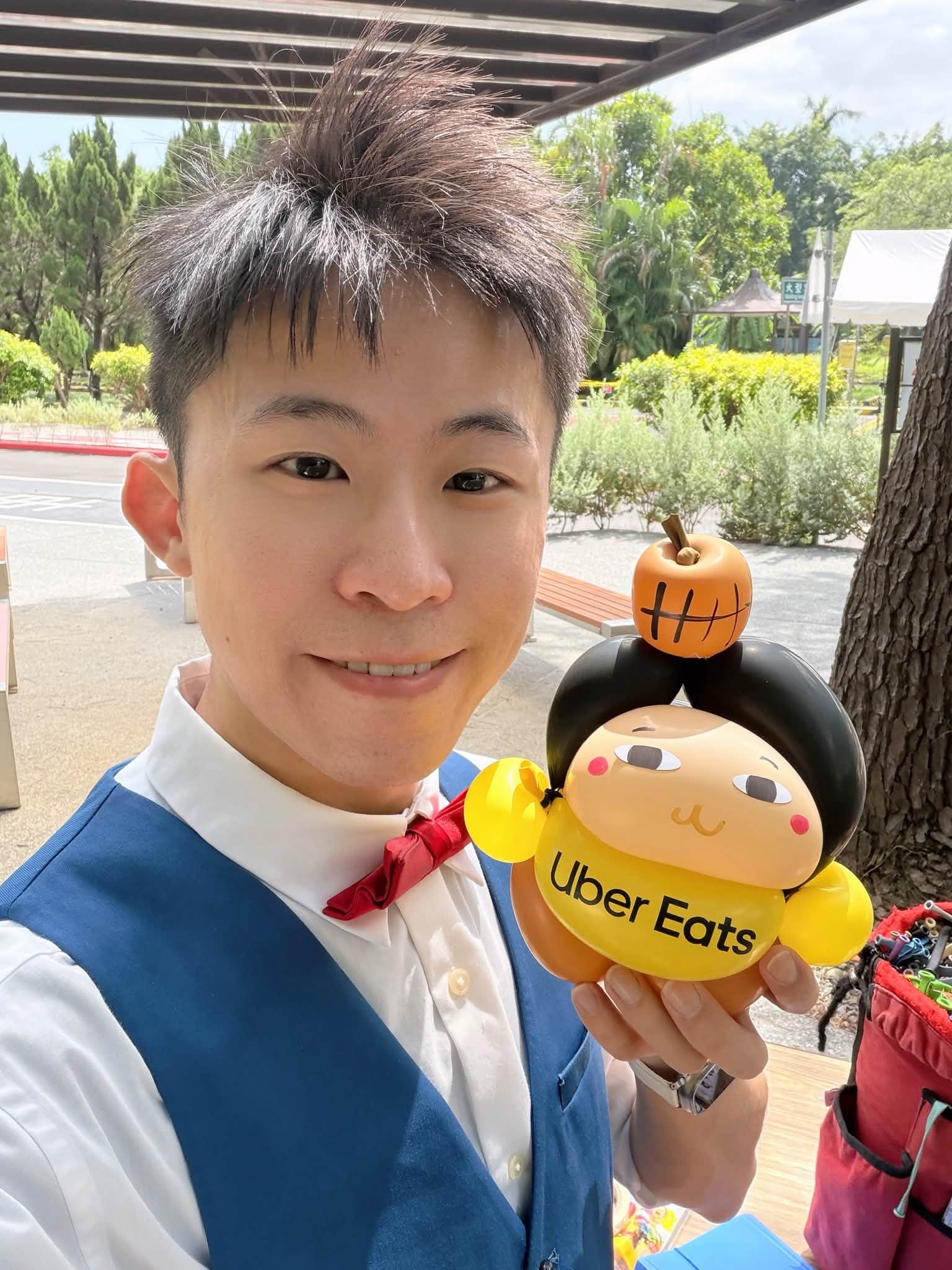 柯男手捧黃色 Uber Eats 圍兜、頭頂橘色小球的娃娃造型氣球自拍，背景是戶外綠樹