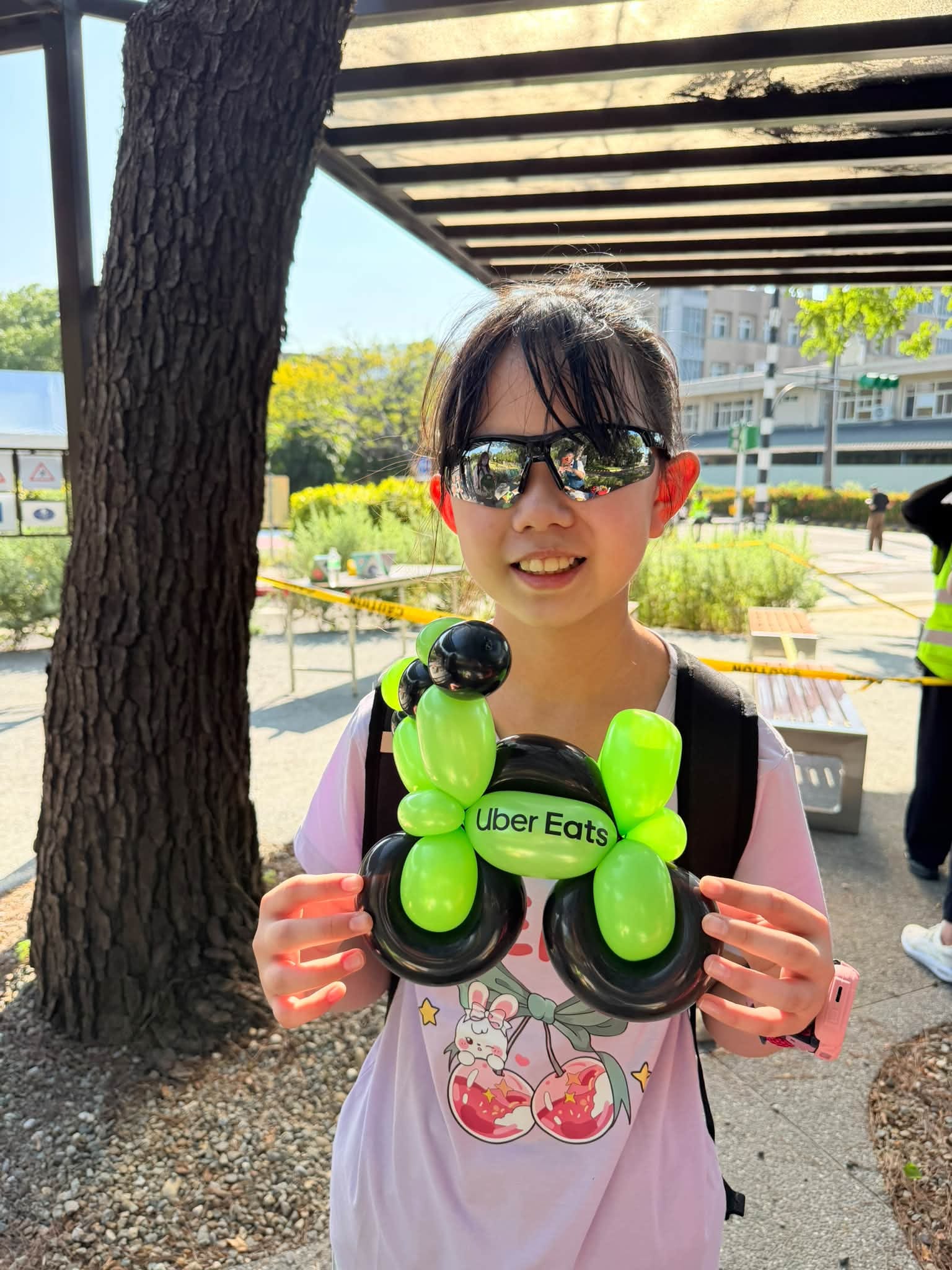 戴墨鏡的女孩雙手拿著綠色車身、黑色輪子的 Uber Eats 機車造型氣球