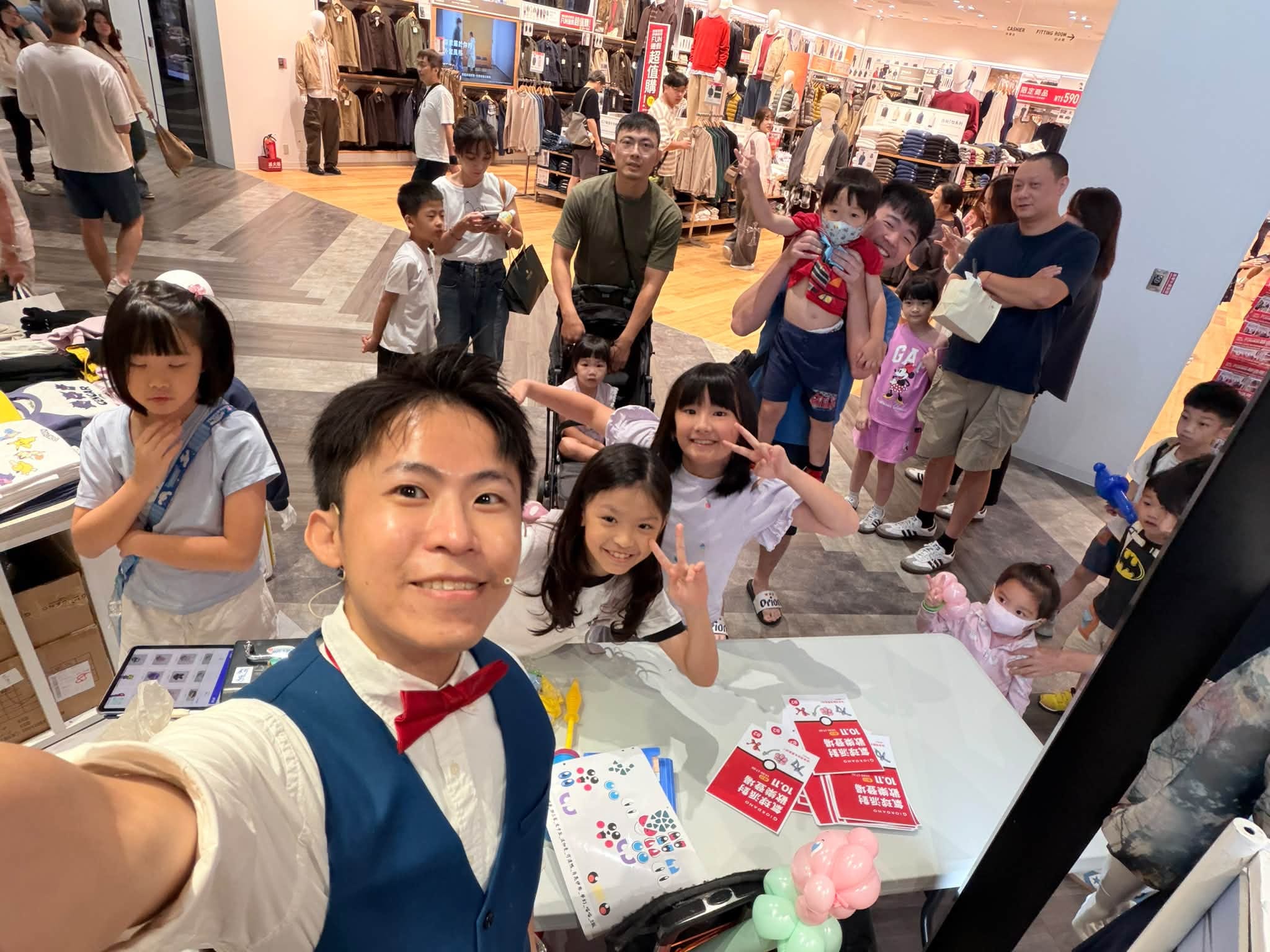 柯男在攤位後方自拍,身後的小女孩們比 YA,家長抱著孩子在後面等候