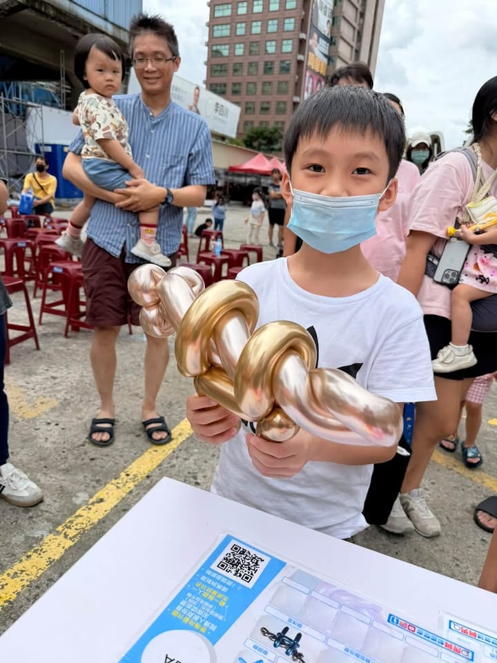 男孩拿著金色的槍造型氣球站在折氣球桌前，後方是觀眾座位區的紅色板凳