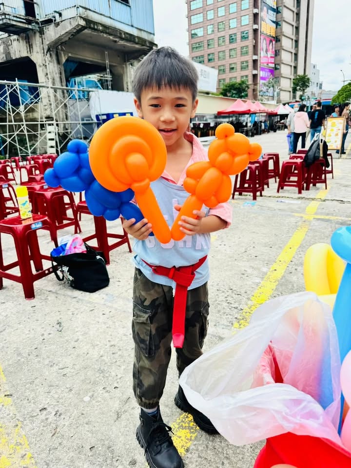 穿迷彩褲的男孩一手拿橘色棒棒糖氣球，一手拿藍色與橘色造型氣球