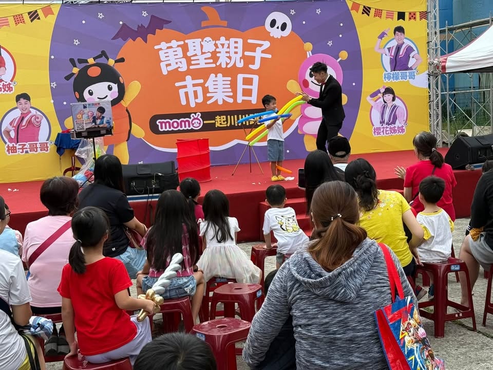 我在舞台上拿著黃藍色長條氣球，跟上台的小男孩互動，台下觀眾坐在紅色板凳上
