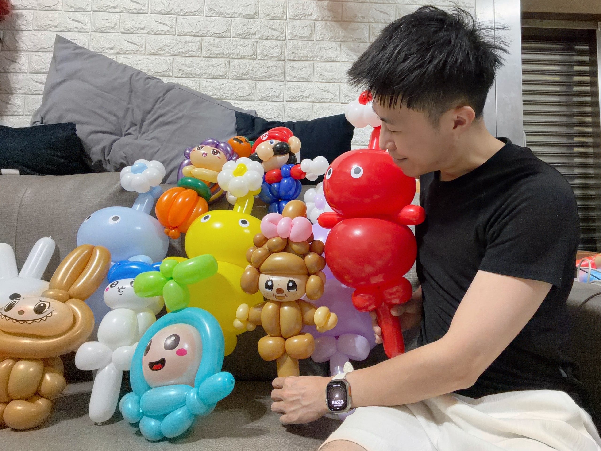 穿黑上衣的男生坐在沙發旁,低頭看著一整排完成的角色造型氣球,手握一隻紅色皮克敏