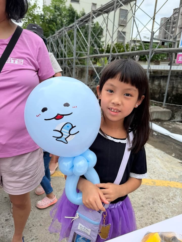 穿紫色紗裙的女孩拿著淺藍色的角落生物蜥蜴造型氣球，吐著舌頭、抱著一條小魚