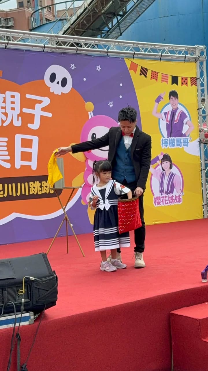 穿水手服的小女孩捧著紅色魔術袋，我拿著黃色絲巾在舞台上表演魔術