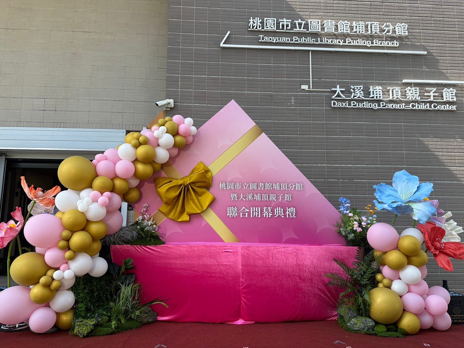金色蝴蝶結的粉色背板前，左側氣球柱沿背板弧形向上，右側氣球柱旁立著藍色與紅色大花