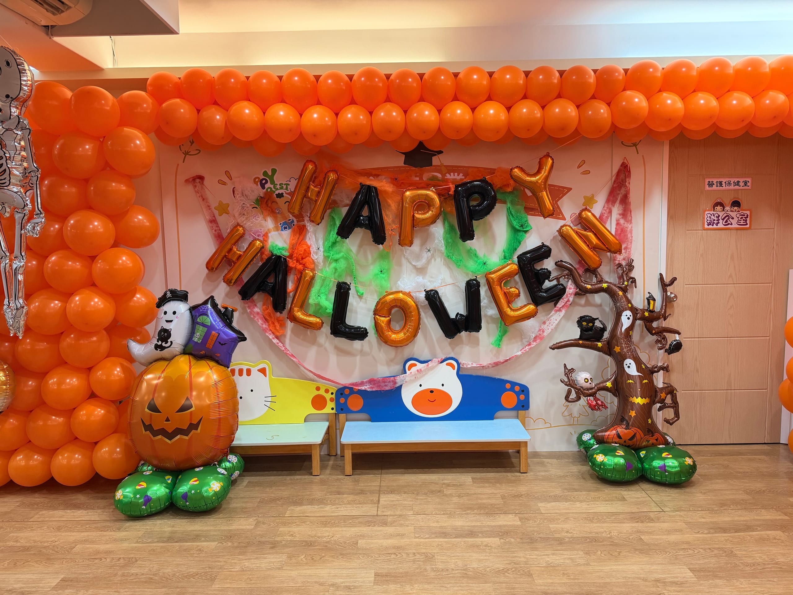 貼著 HAPPY HALLOWEEN 字樣的背板，兩側是橘色氣球牆，前方有南瓜與鬼魂造型氣球和一個樹造型裝飾