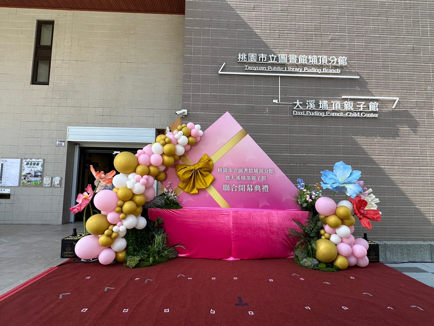 粉色禮物盒背板左右兩側的金色、粉色、白色氣球柱，搭配大朵紙花與綠色植栽，下方鋪紅地毯