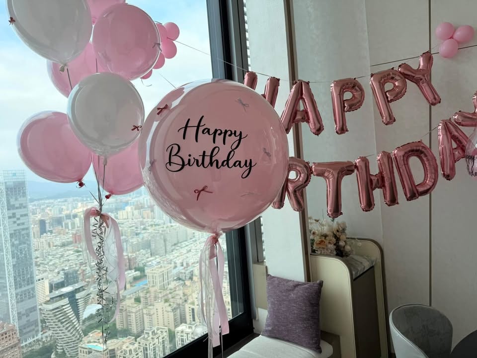 窗邊的粉色與白色氣球束，主球是貼蝴蝶結的 Happy Birthday 雙層氣球，背景是高樓城市景觀