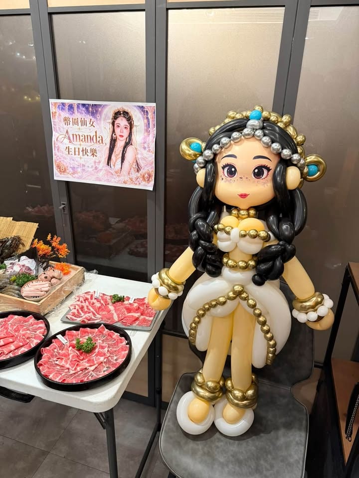 白金禮服氣球人偶坐在椅子上，後方牆上貼著生日海報，旁邊桌上擺著火鍋肉盤