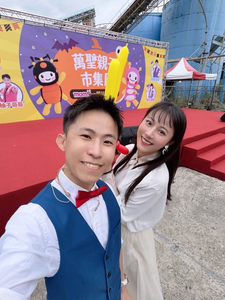 穿藍色背心、紅色領結的我在萬聖節主題舞台前自拍，身旁是穿白衣的長髮女主持人