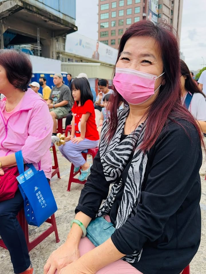 我媽坐在台下紅色板凳上，戴粉紅色口罩、圍斑馬紋圍巾，笑得眼睛瞇起來