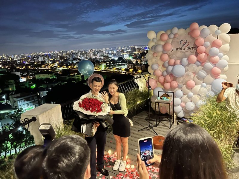 求婚成功的新人捧著紅玫瑰花束在粉藍氣球牆與台中夜景前合照